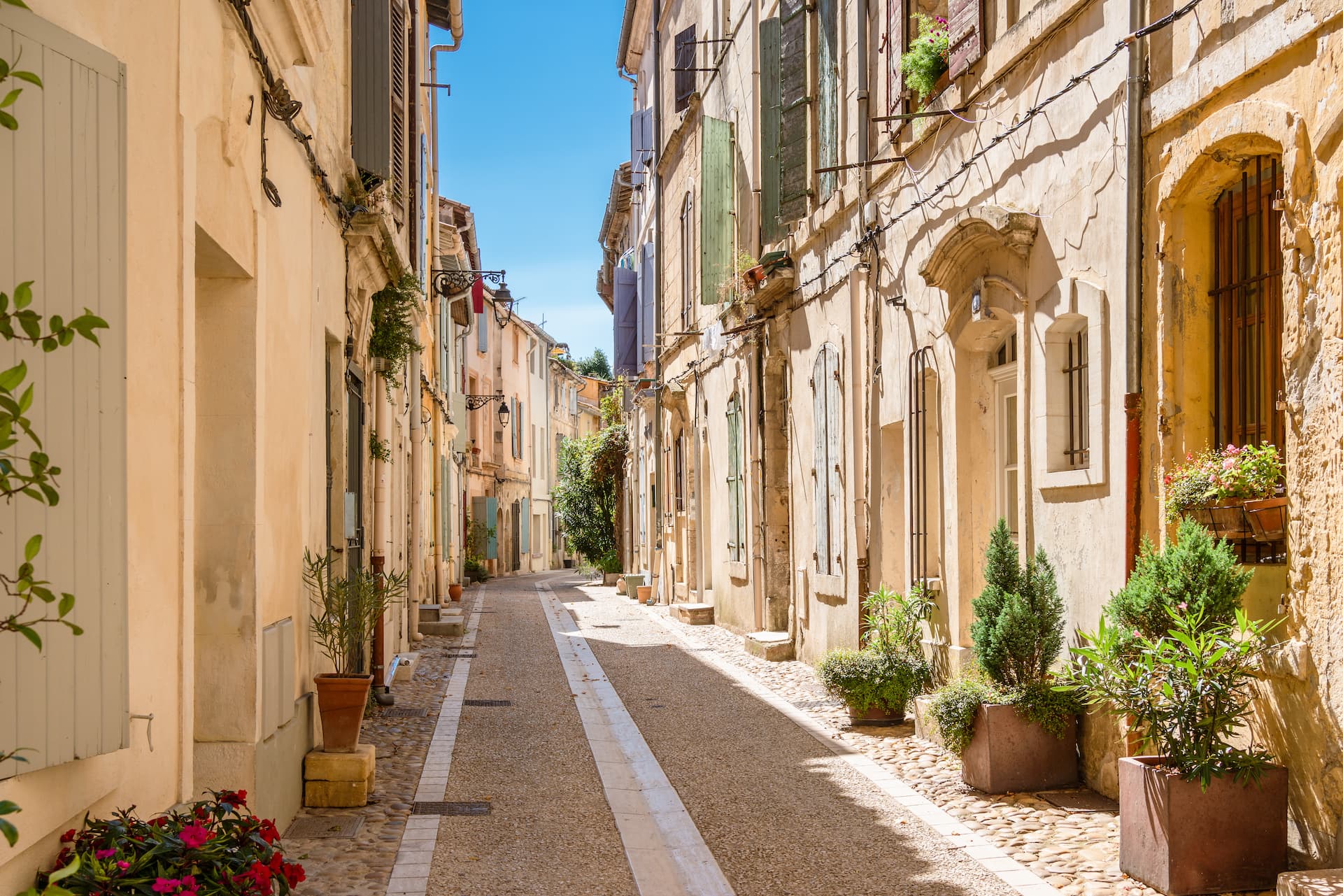 Narrow cobblestone street lined with historic stone buildings and potted plants under a clear blue sky in Arles.