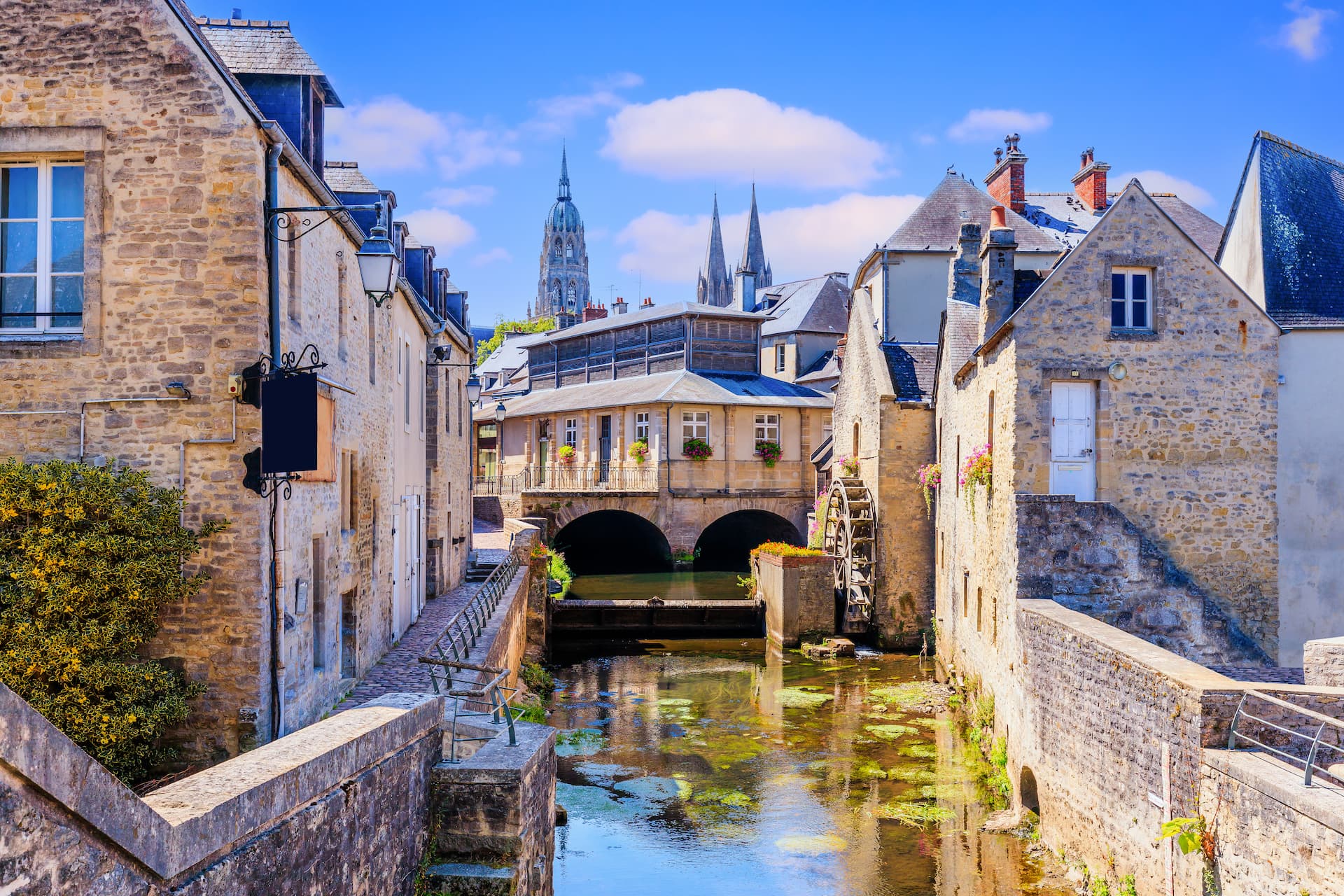 Stone buildings line a narrow river with a waterwheel, spires visible in the distance.