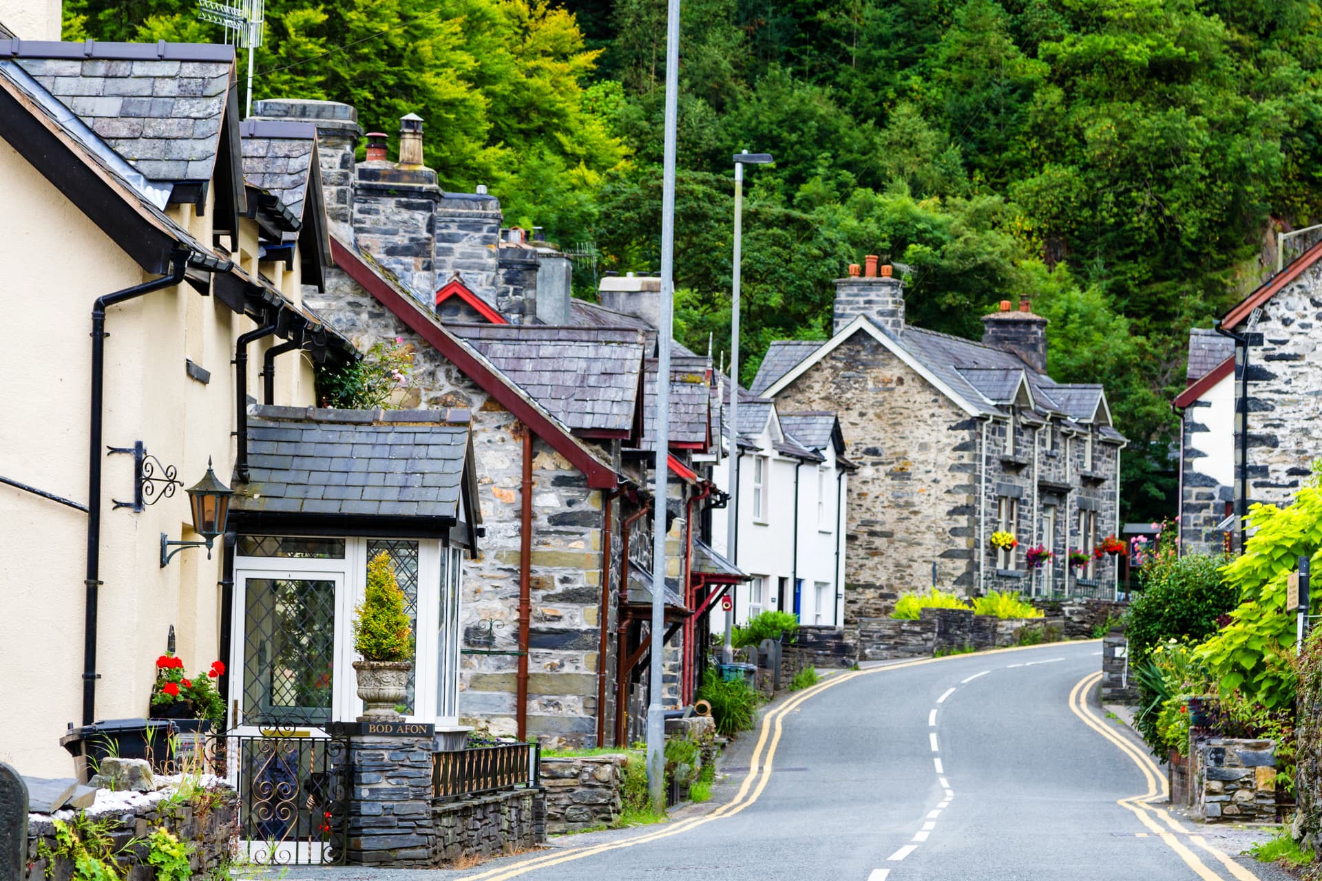Curving street lined with stone houses and lush green forest in Betws-y-Coed.