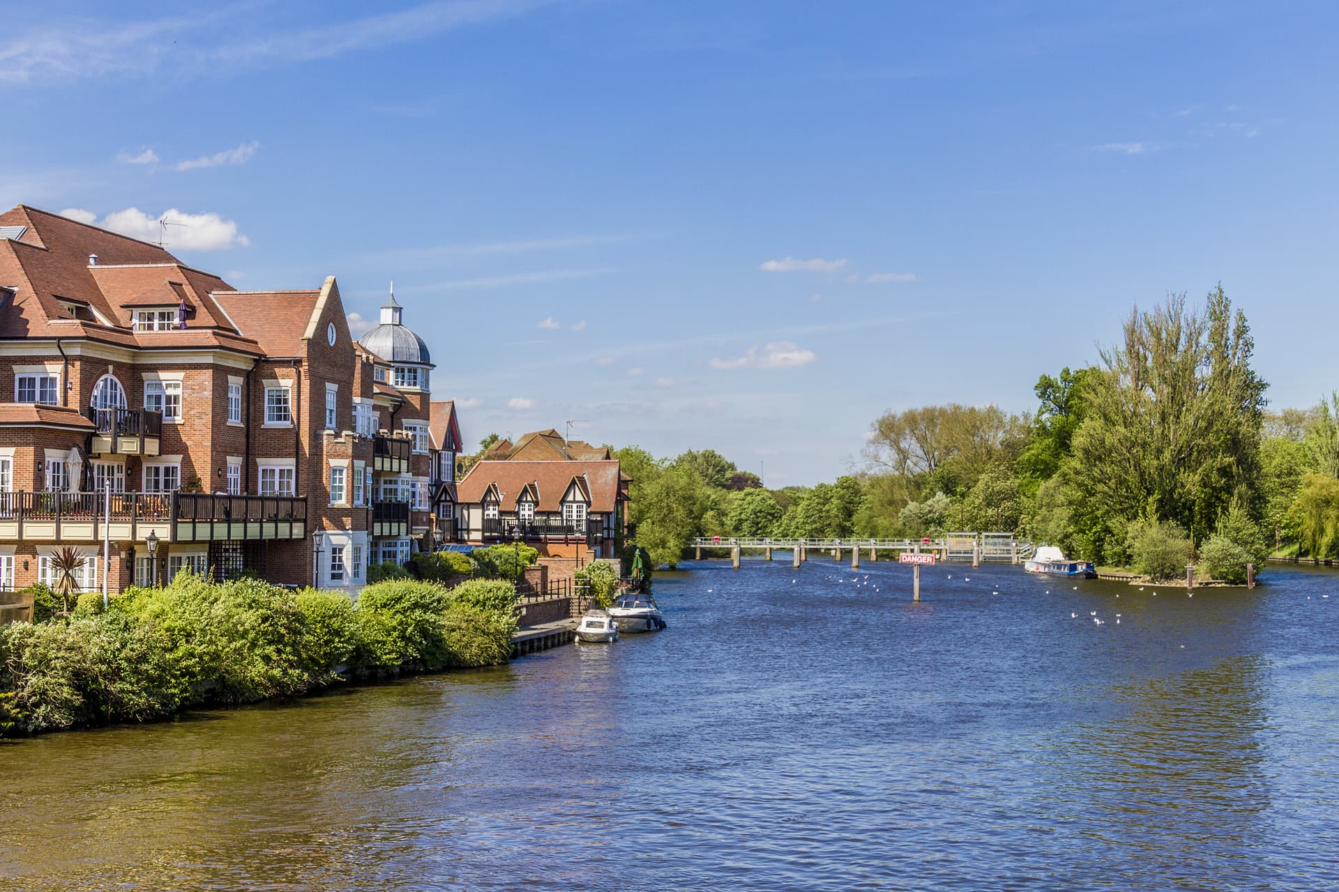 Brick buildings along a riverbank with boats moored under a bright blue sky in Windsor Town.