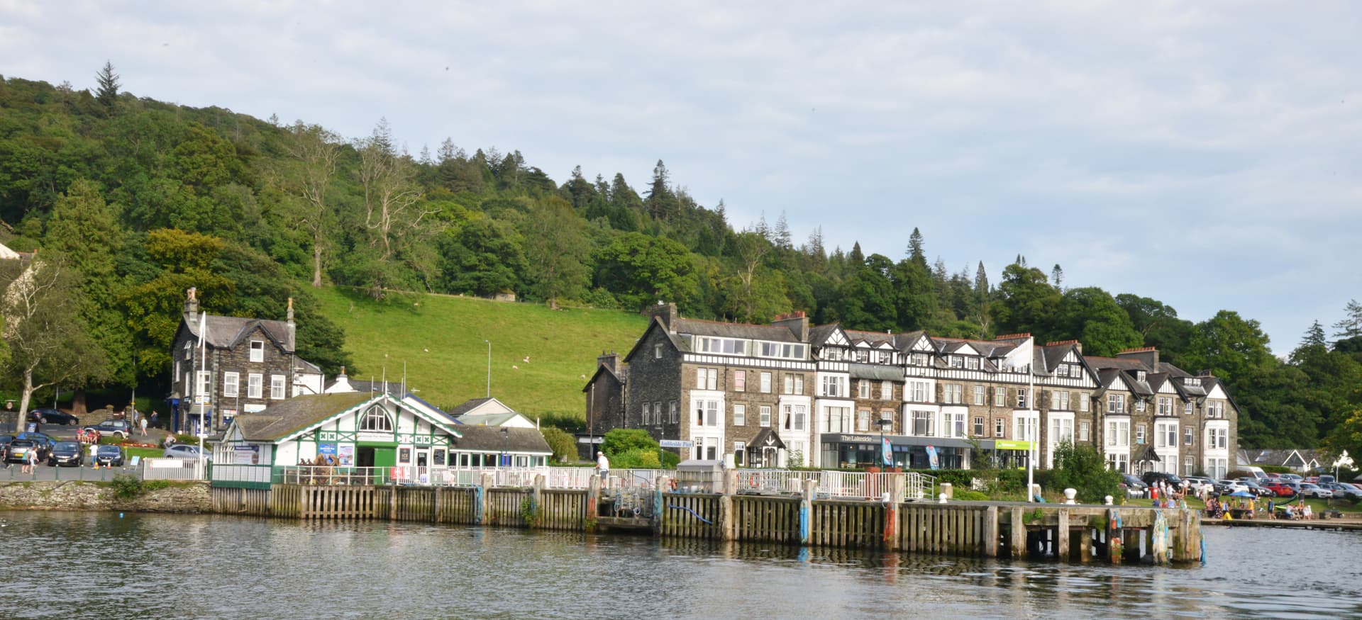 Ambleside Pier Waterhead buildings by lake with wooded hillside and parked cars.