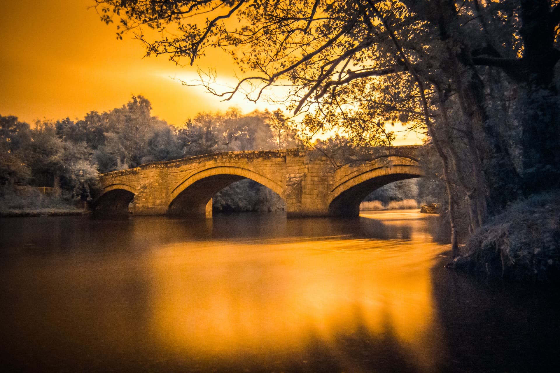 Stone arch bridge over river at sunset with orange sky and silhouetted trees, Pooley Bridge.