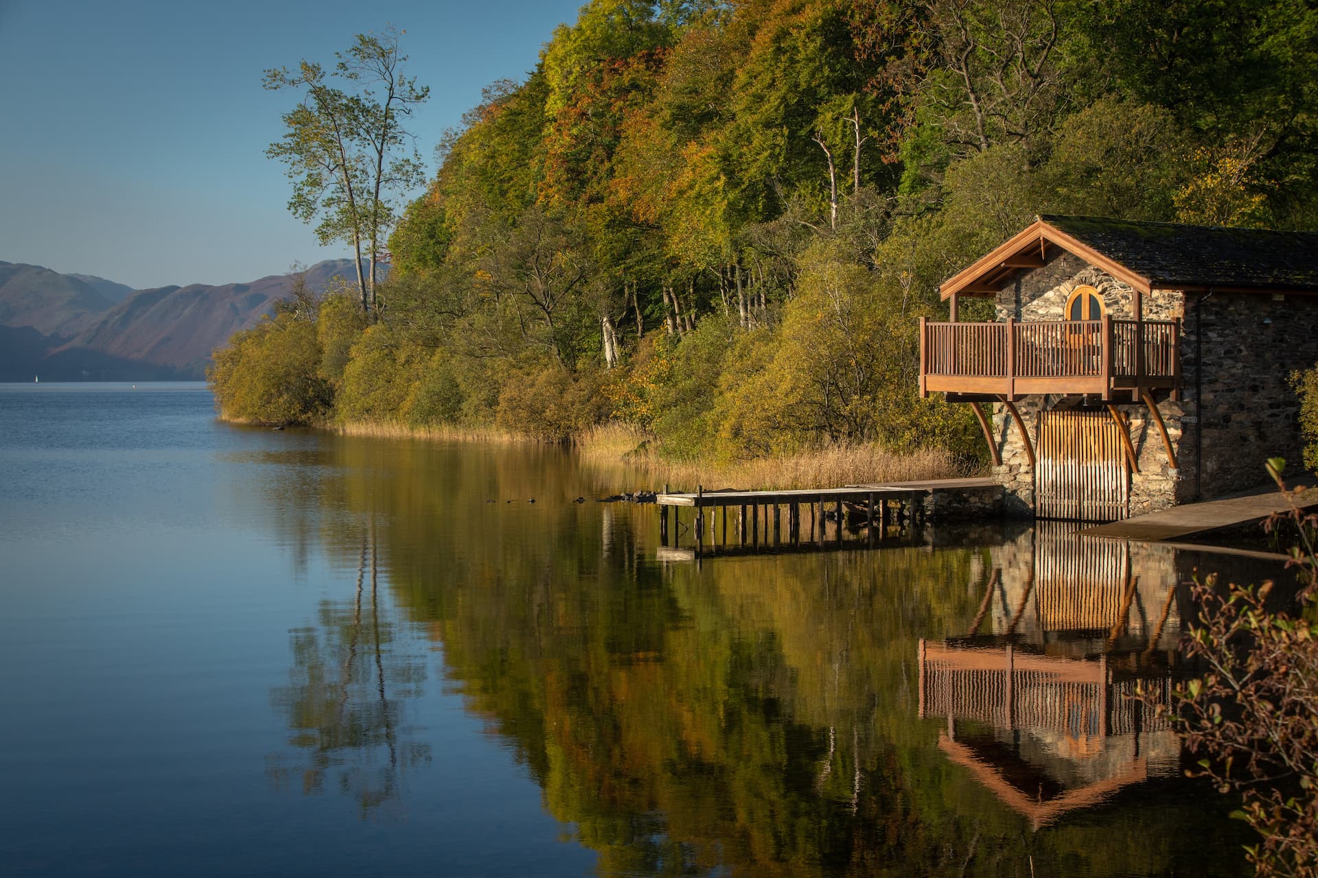 Stone boathouse with wooden balcony on Ullswater lake with autumn foliage and mountains.