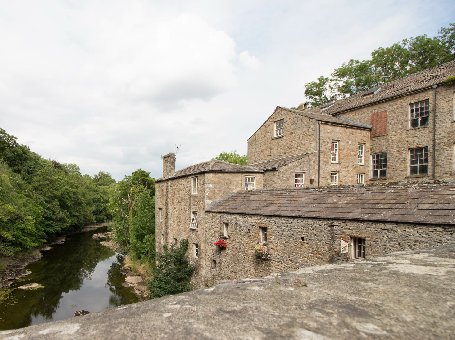 Stone mill buildings beside a river surrounded by dense green trees under a cloudy sky, Aysgarth.