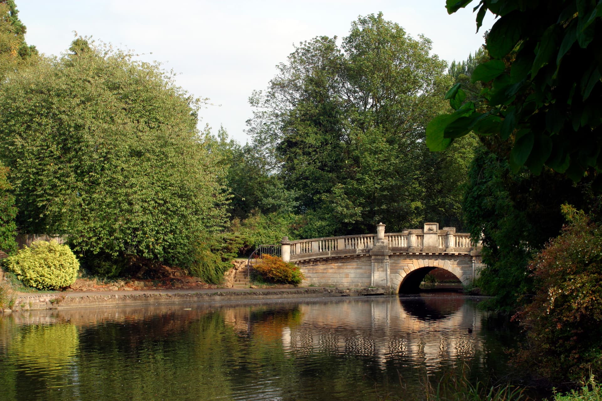Stone bridge over water reflecting lush green trees in Pittville Park, Cheltenham.