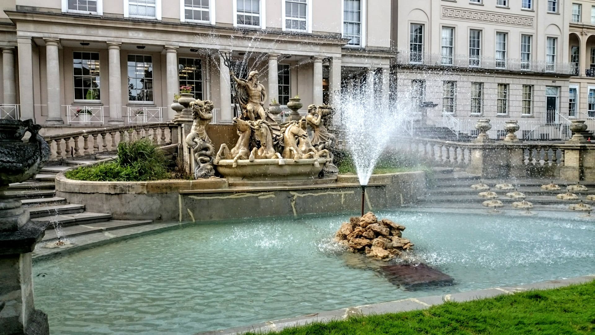 Neptune fountain with statues and splashing water in front of a classical building in Cheltenham.