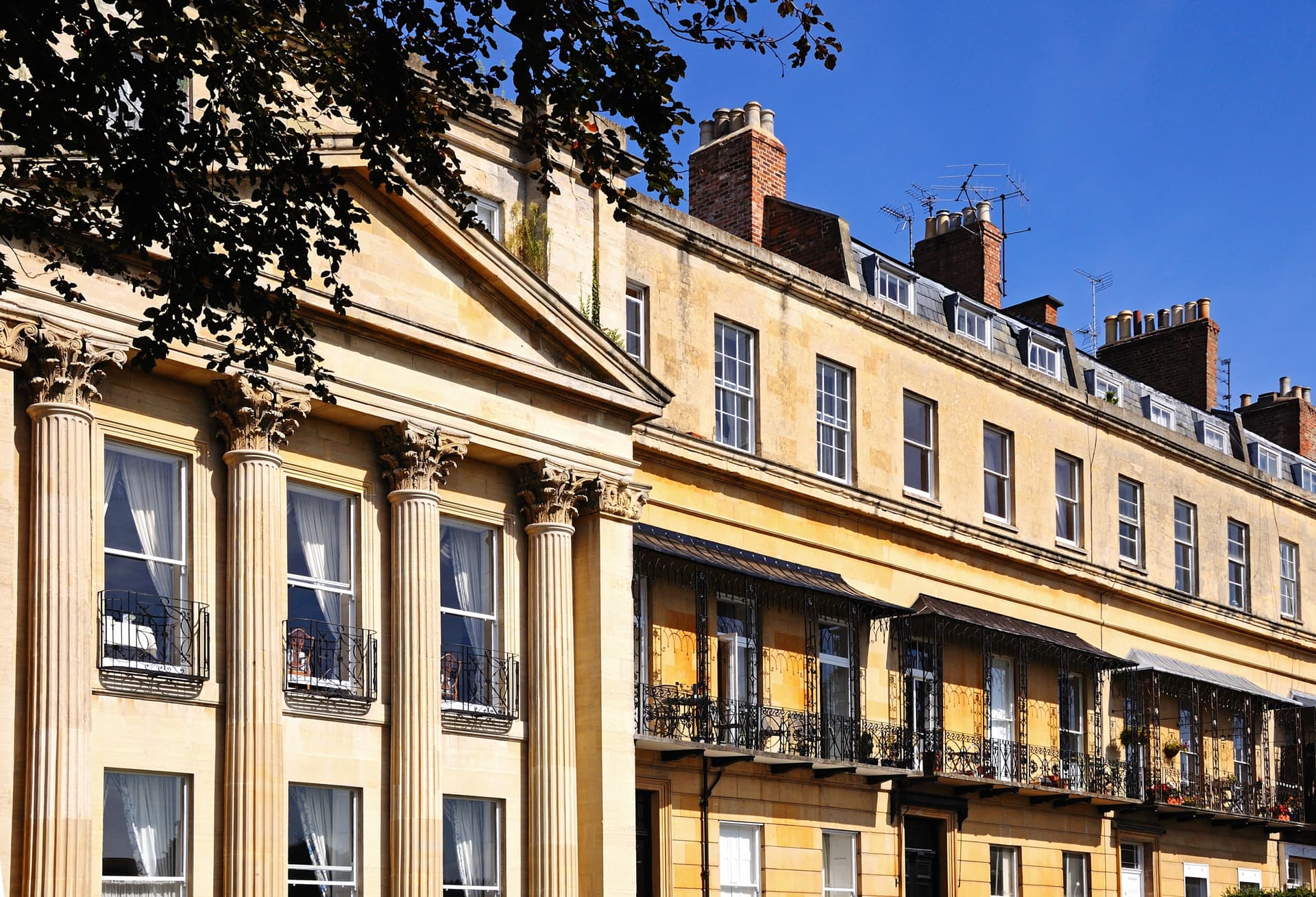 Georgian terraced buildings with stone facades, iron balconies, and chimneys under a clear blue sky in Cheltenham.