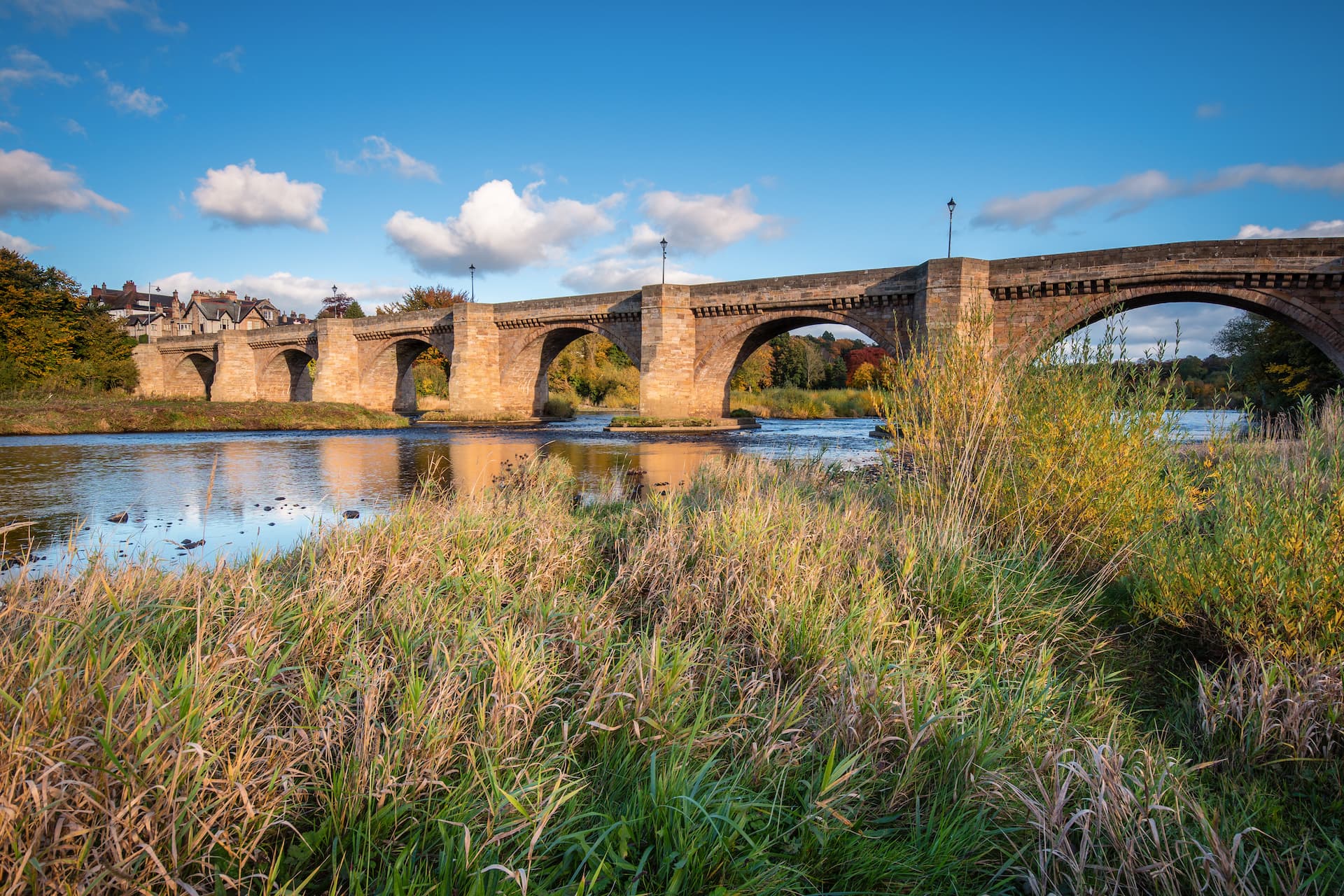 Stone arch bridge over river with autumn foliage and houses on bank, Corbridge.
