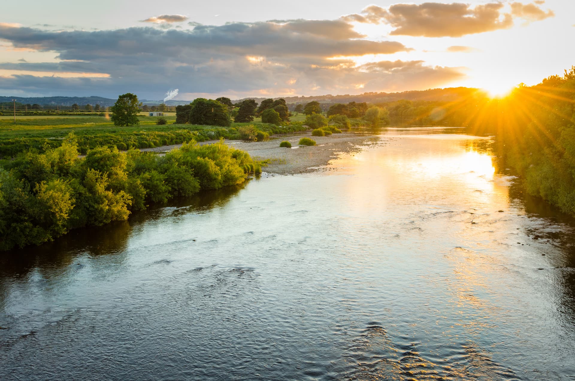 River Tyne at sunset with bright sun flares over green banks and rolling hills in Corbridge.