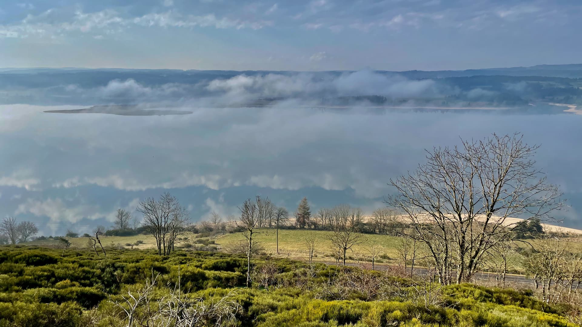 Lake Langogne in spring with low fog over the water and bare trees on the foreground hillside.