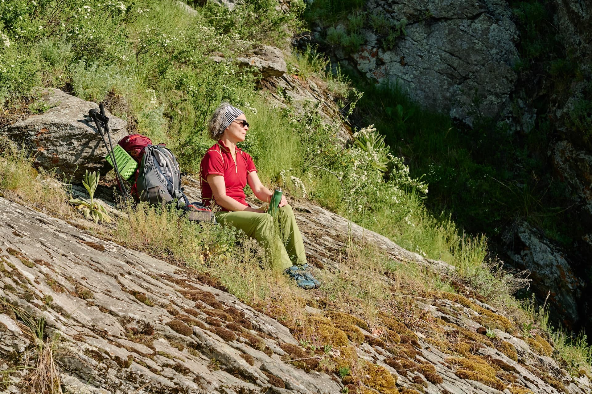 Hiker resting on mossy rocks with backpack and trekking poles on a grassy mountainside.