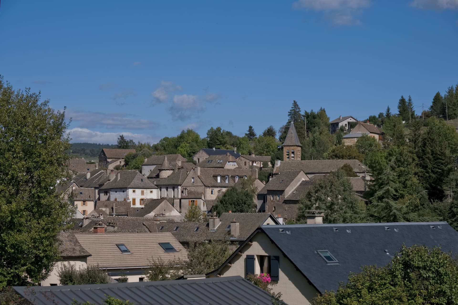 Rooftops of stone houses in Le Bleymard village with a church spire and green trees under blue sky.
