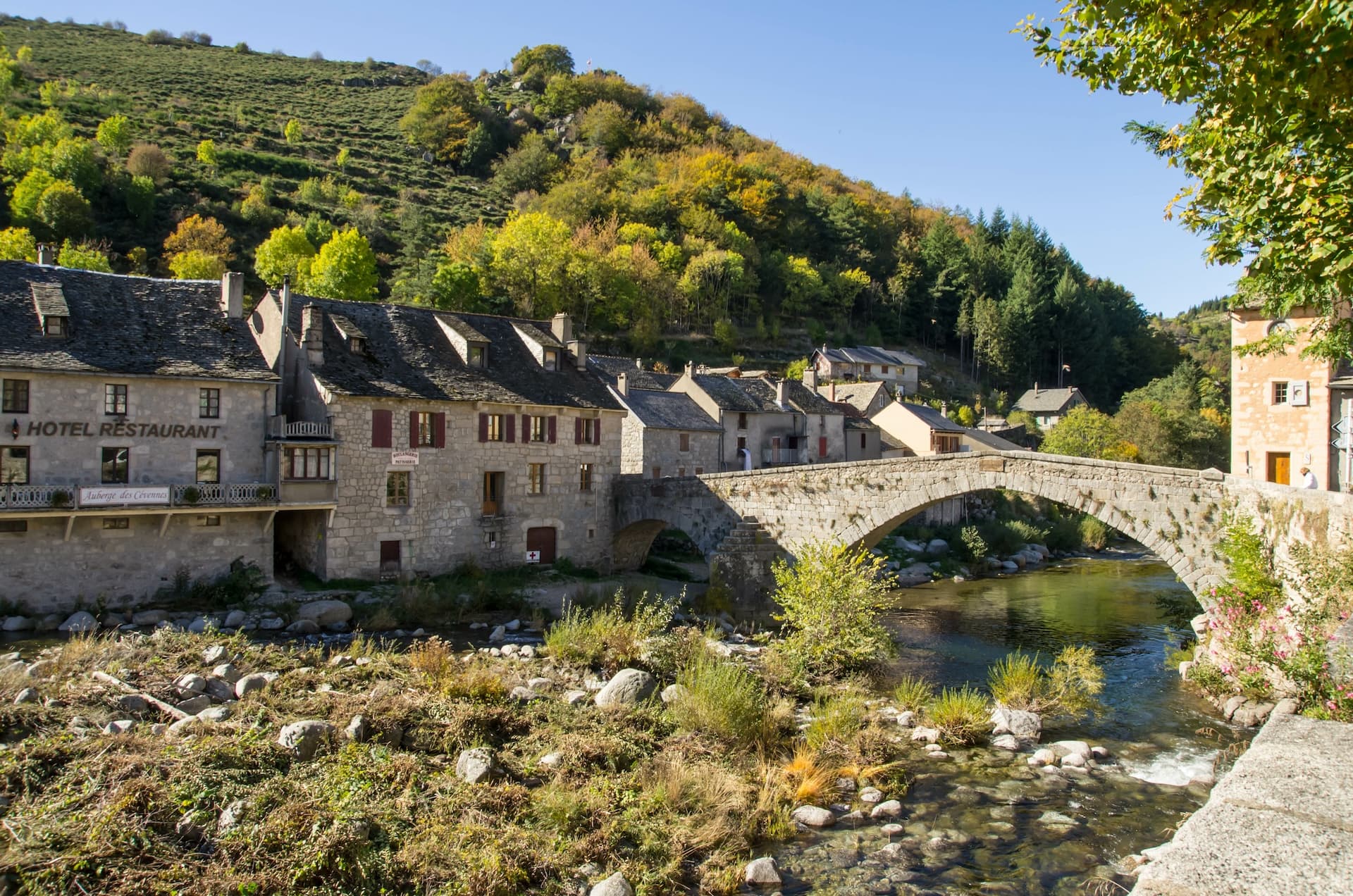 Stone bridge over river in Pont de Montvert village with stone buildings and autumn hillside.
