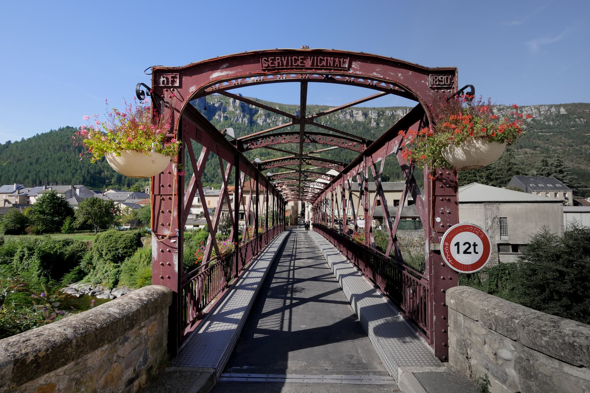 Red metal truss bridge with "Service Vicinal 1890" arch over a village in Florac, France.