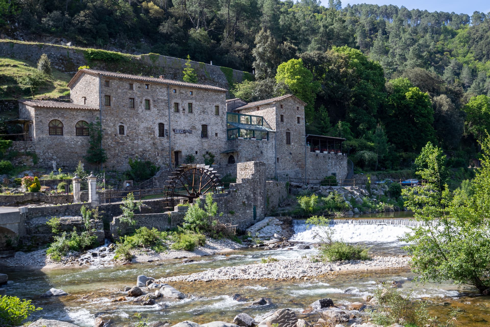 Stone watermill building next to a river with a waterwheel, set against a forested hillside.