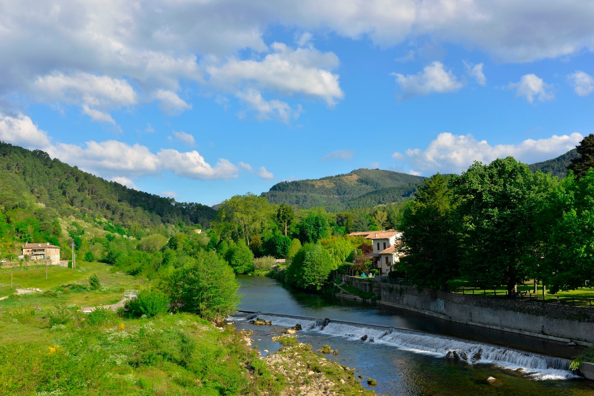 River with small weir flowing through green valley with forested hills in Saint-Jean-du-Gard.