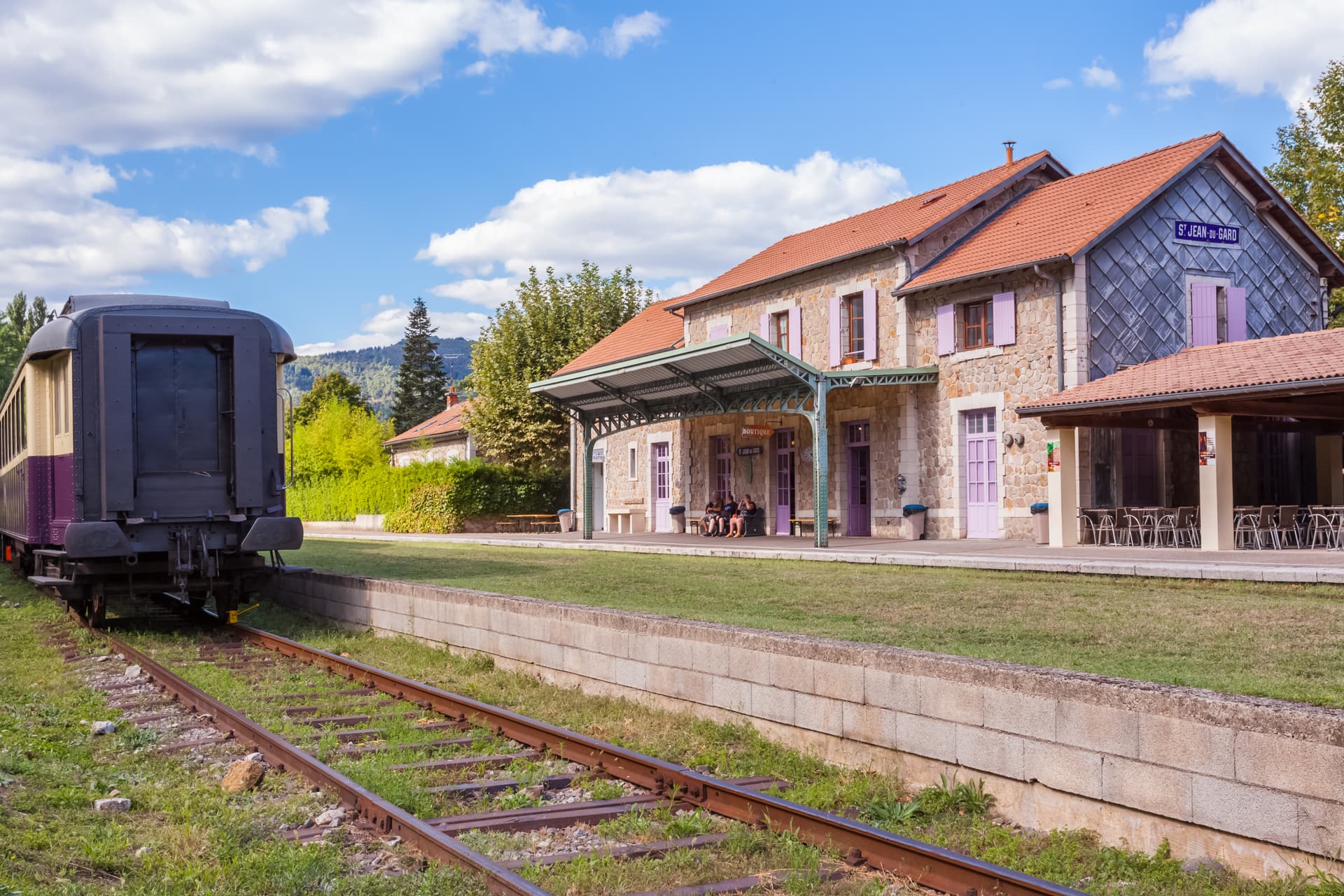 Train car parked next to stone station building in Saint-Jean-du-Gard under blue sky.