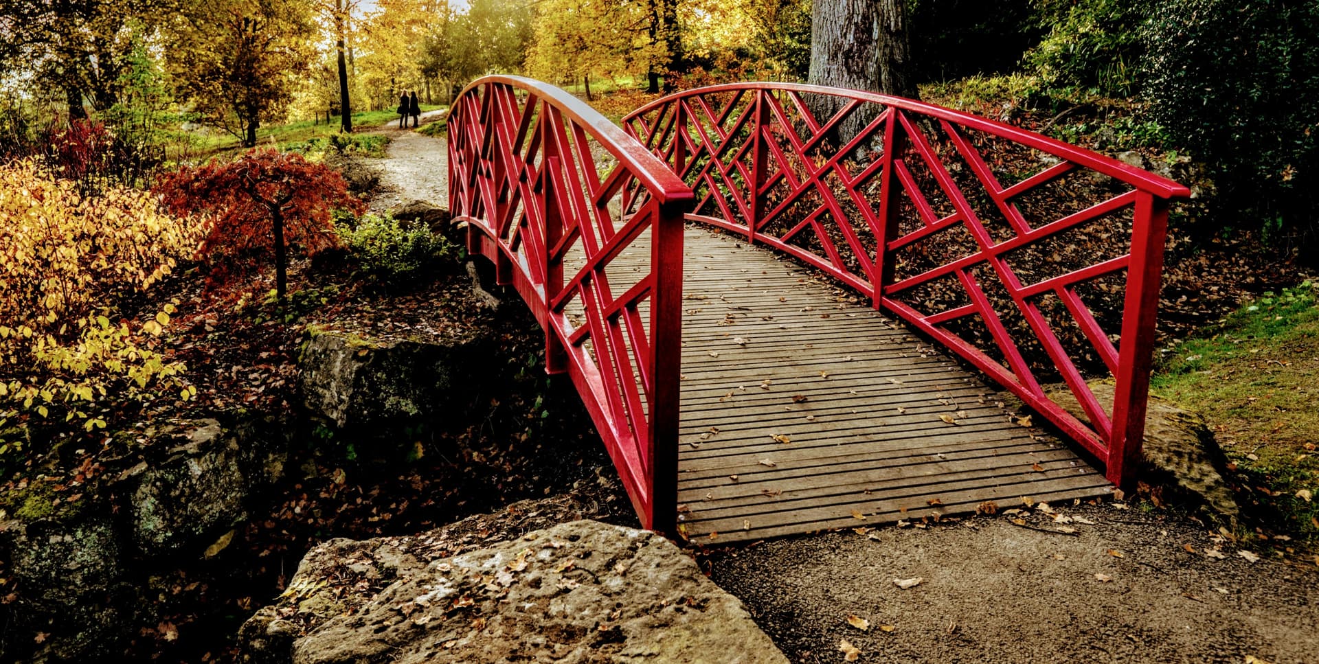 Red arched bridge in Batsford Arboretum with autumn foliage and path leading away.