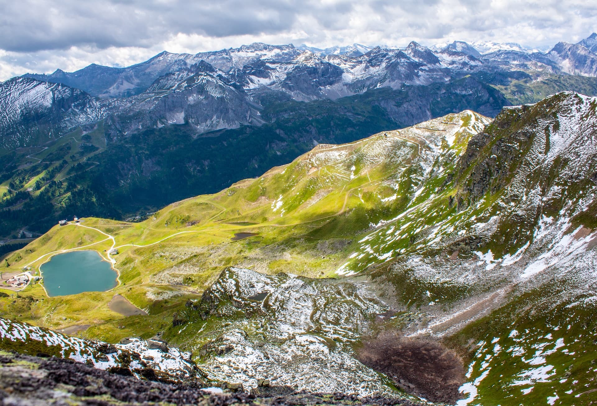 Alpine lake below snow-dusted green slopes with rugged, snow-capped mountains in the background.