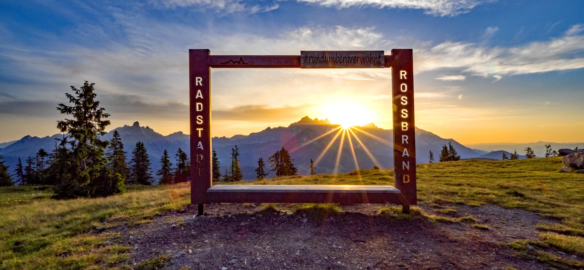 Wooden viewpoint frame at Rossbrand summit during sunset with mountain range backdrop.
