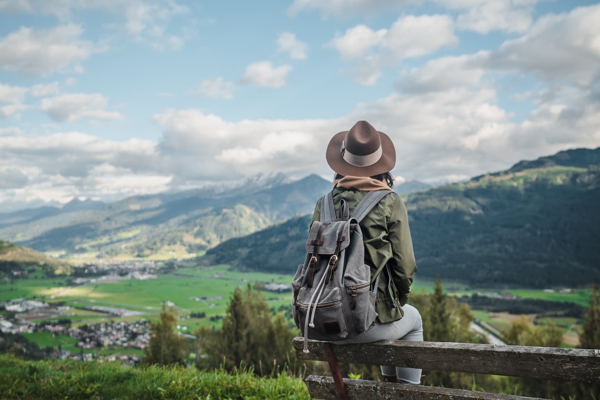 Hiker with backpack and hat resting on bench overlooking Austrian alpine valley and mountains.