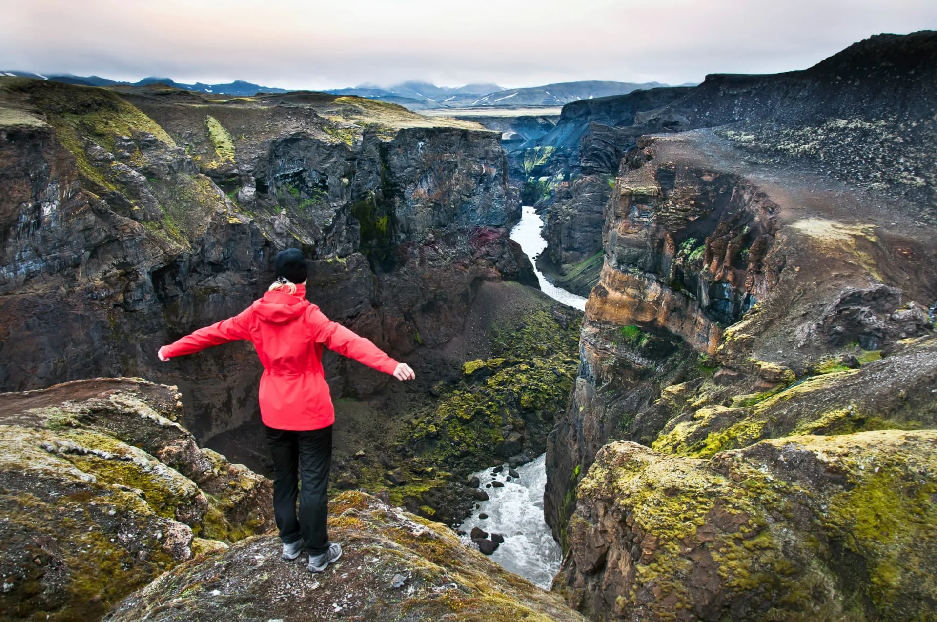 Hiker in red jacket overlooking Markarfljotsgljufur Canyon with river and distant mountains.