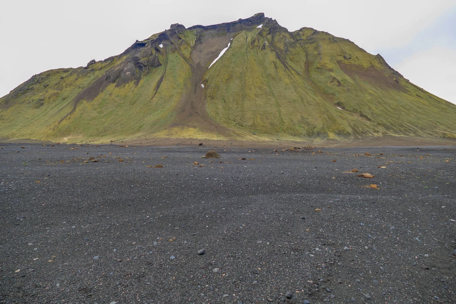 Hiking toward Hrafntinnusker Hut across black volcanic gravel toward mossy green mountain.