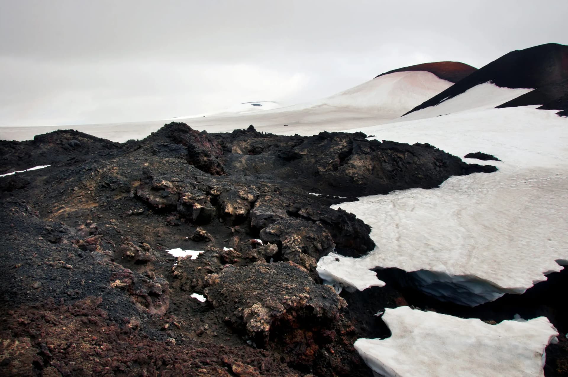 Lava field terrain with snow patches and dark volcanic slopes under an overcast sky in Fimmvörðuháls area.