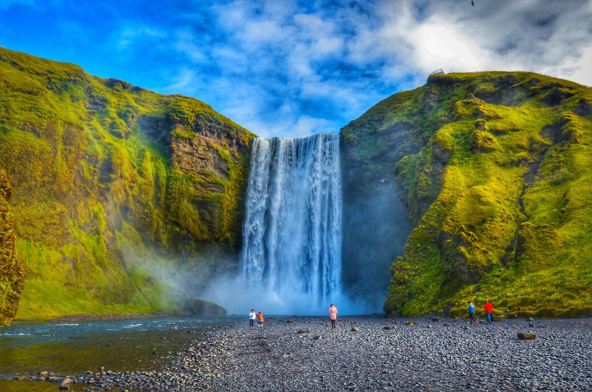 Skogafoss waterfall cascading between mossy green cliffs with tourists on the rocky riverbank.