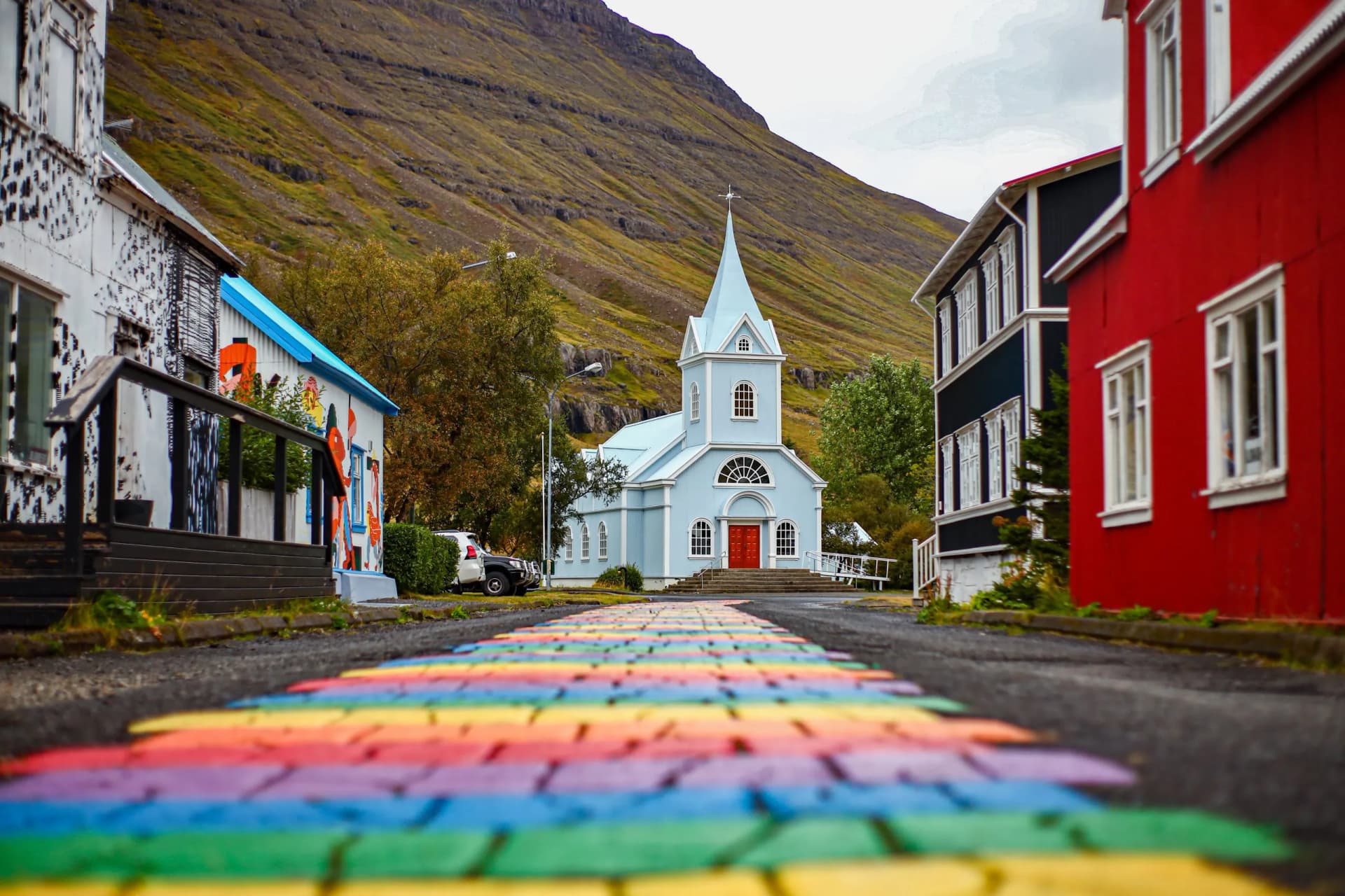 Light blue church in Seydisfjordur with rainbow path leading toward steep, grassy mountainside.