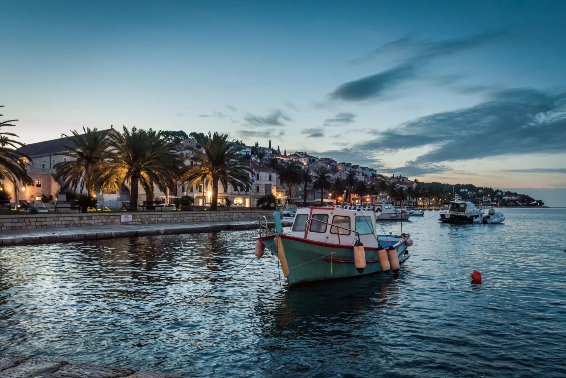 Small boat moored in Hvar harbour at dusk with illuminated palm trees and hillside town.