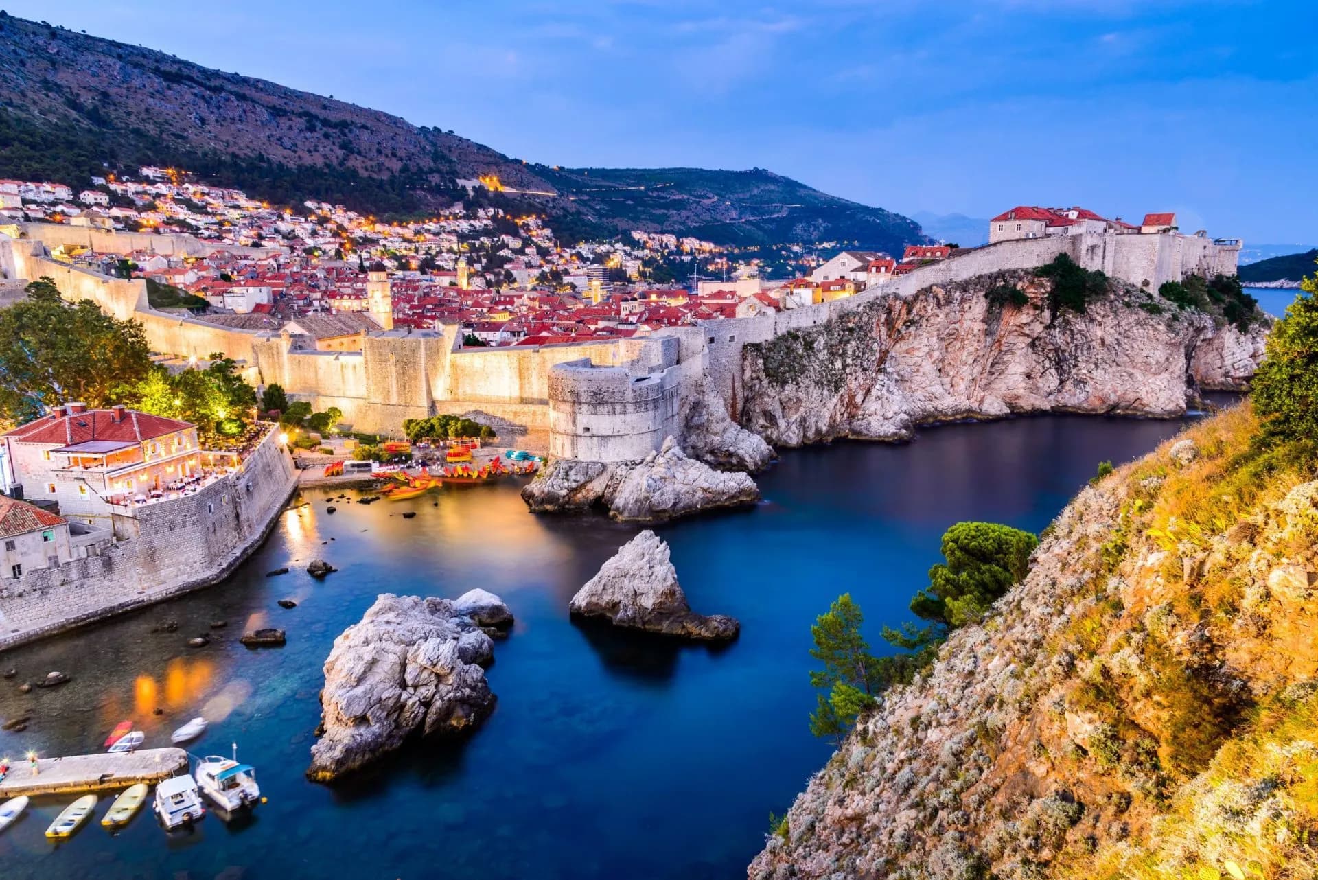 Dubrovnik, Croatia illuminated old town walls and harbor at twilight with boats on dark blue water.