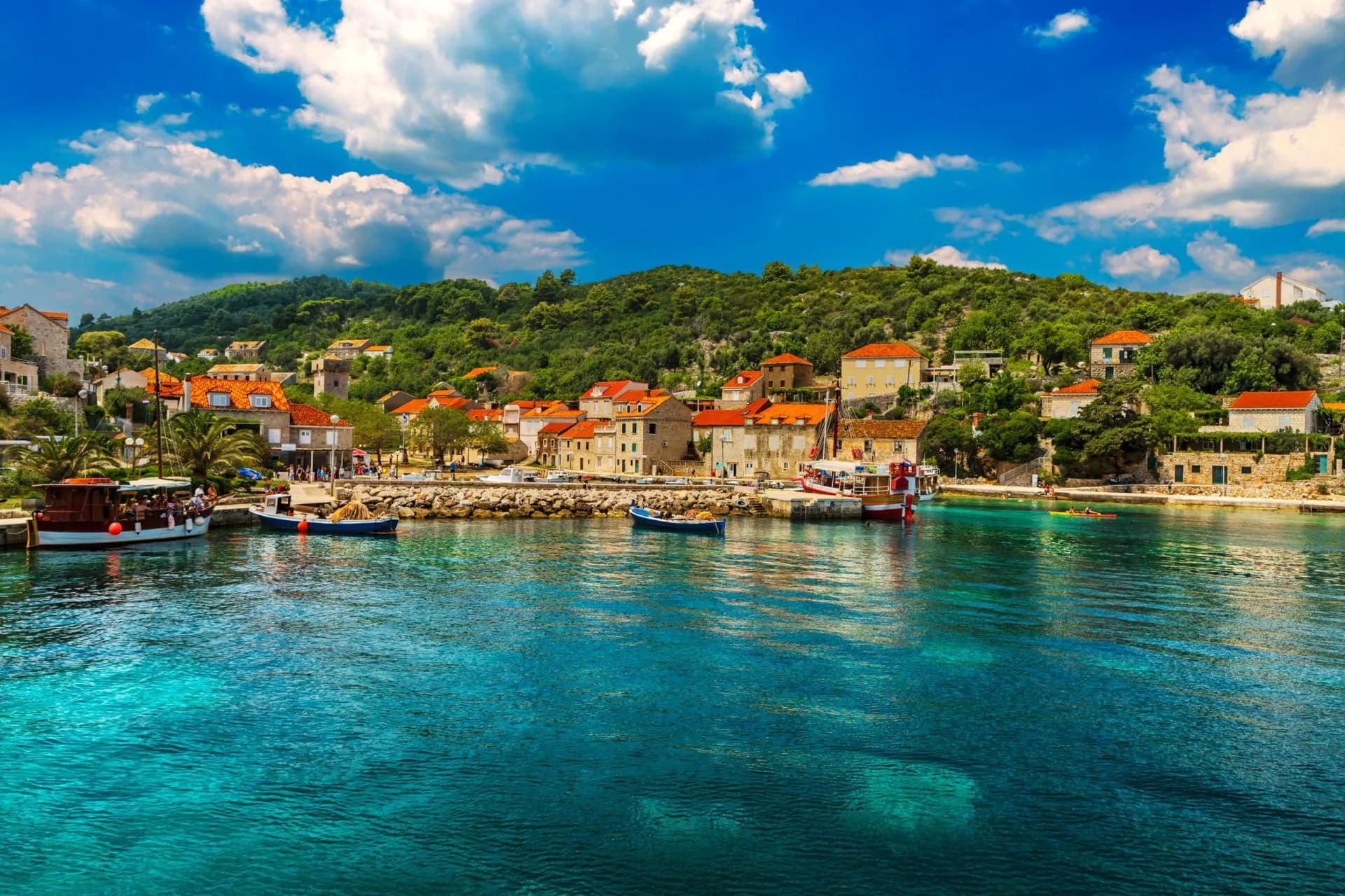 Boats docked in harbor with turquoise water, coastal village, and lush green hillside under blue sky.