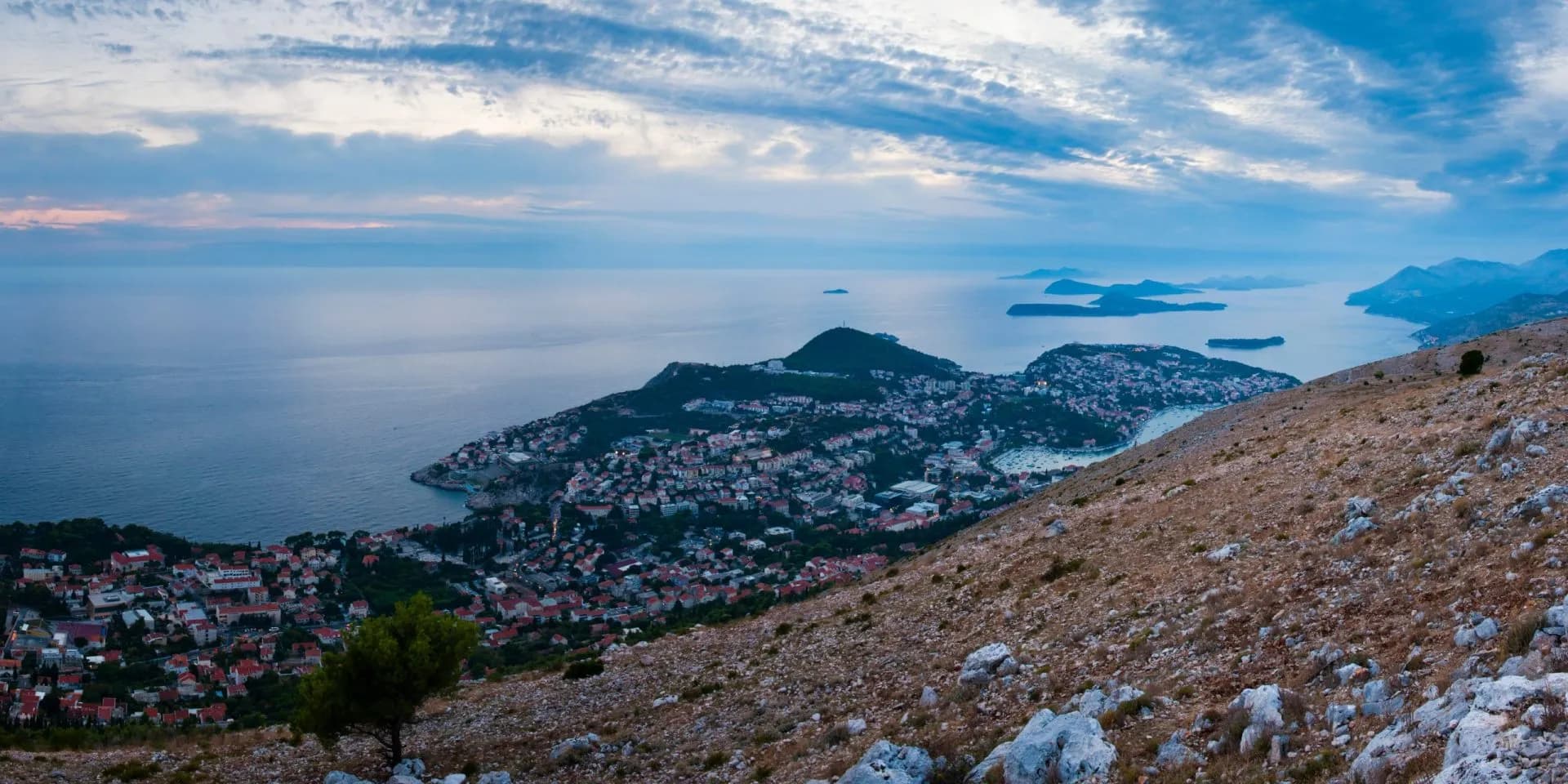 Coastal town nestled by the sea, viewed from a rocky, arid hillside under a cloudy sky.