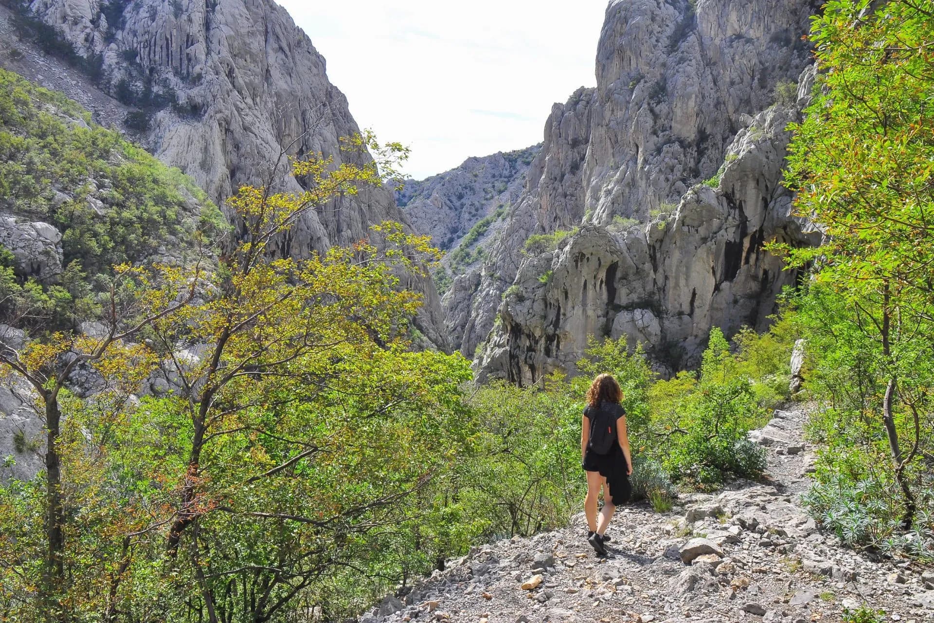 Hiker walking on rocky trail through Paklenica canyon with steep limestone cliffs and green foliage.