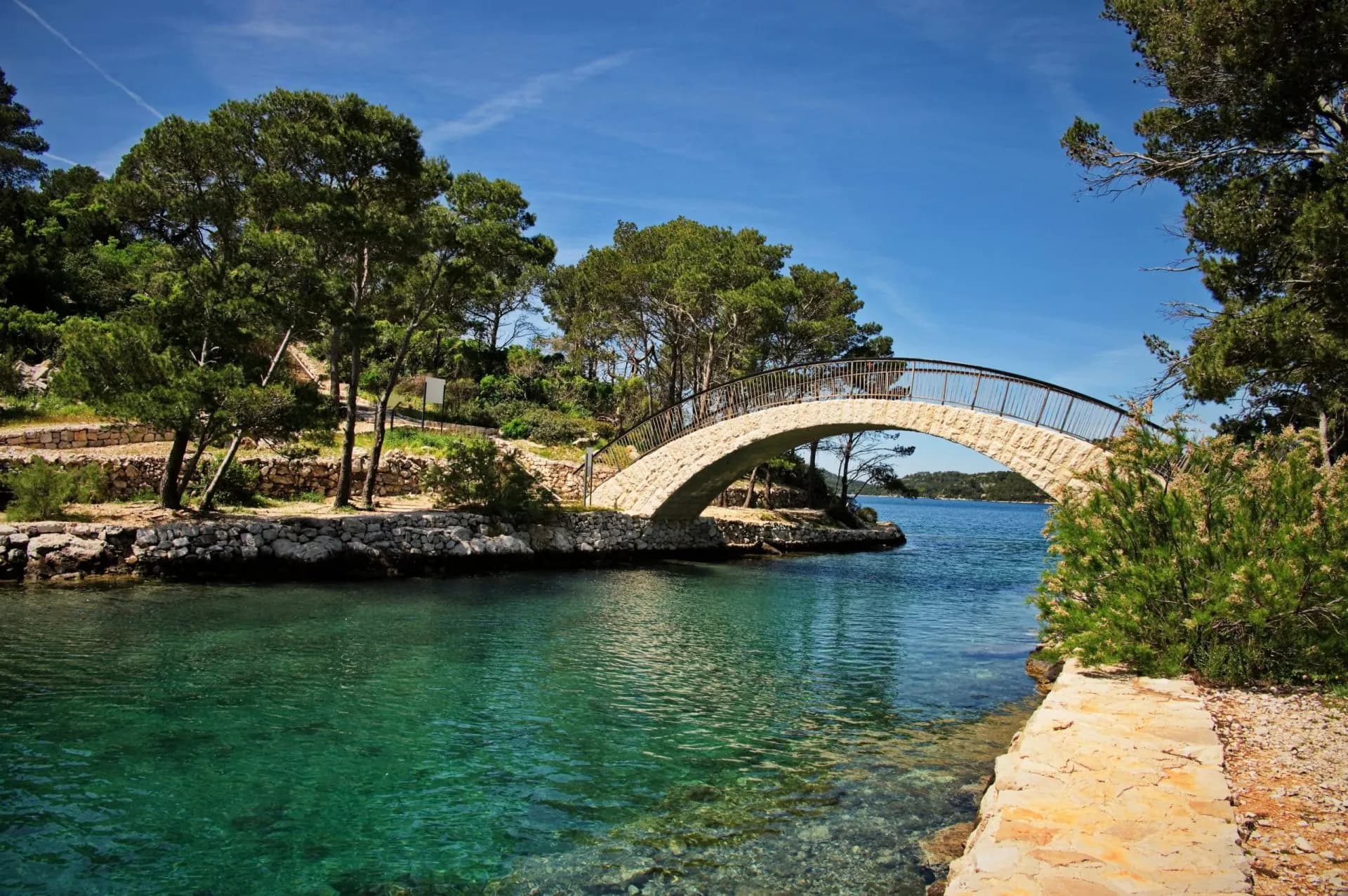 Stone arch bridge over turquoise water on Mljet Island, surrounded by green trees under blue sky.