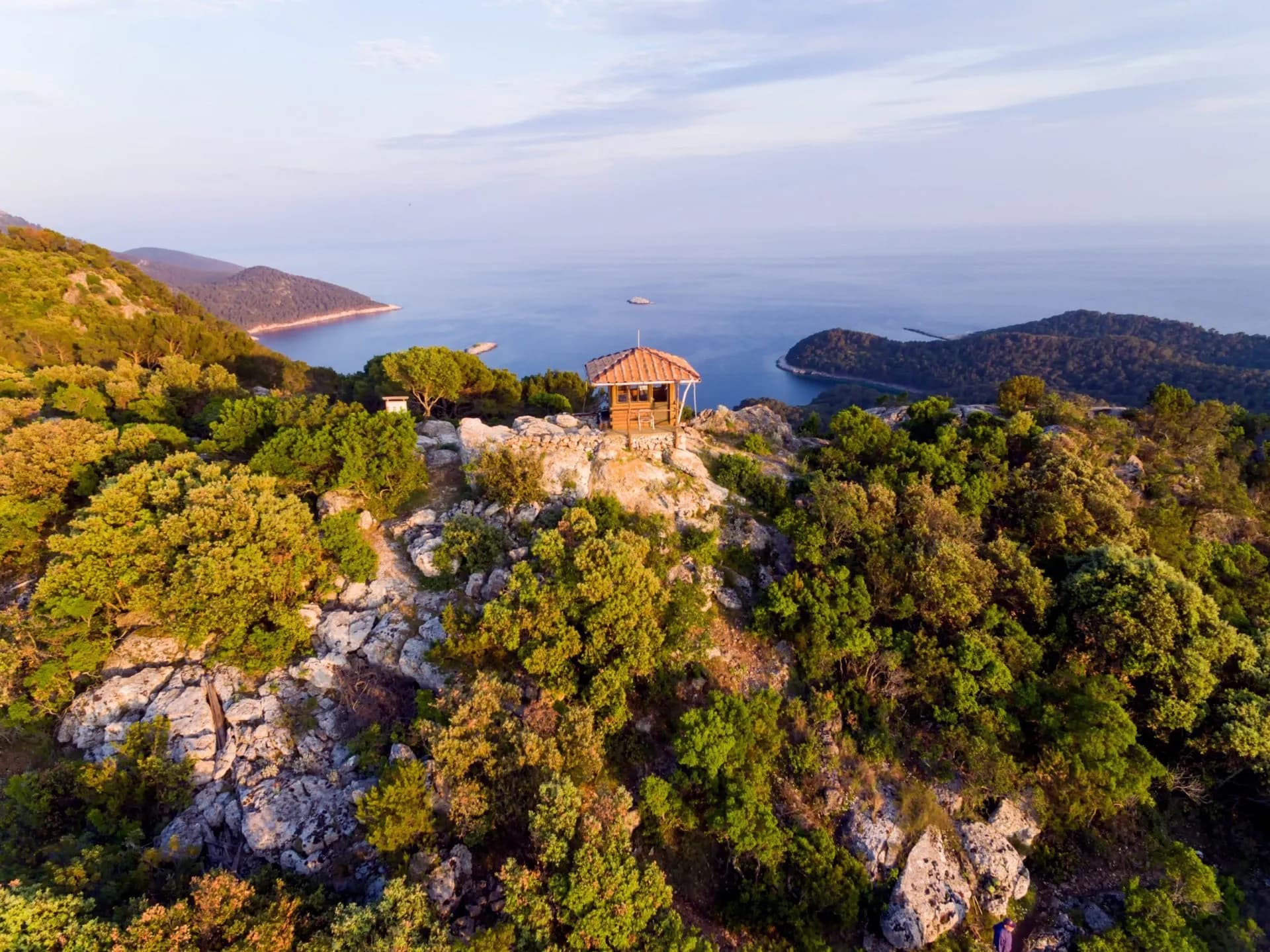 Small wooden viewpoint structure on rocky, forested hill overlooking calm blue sea and distant coastline.