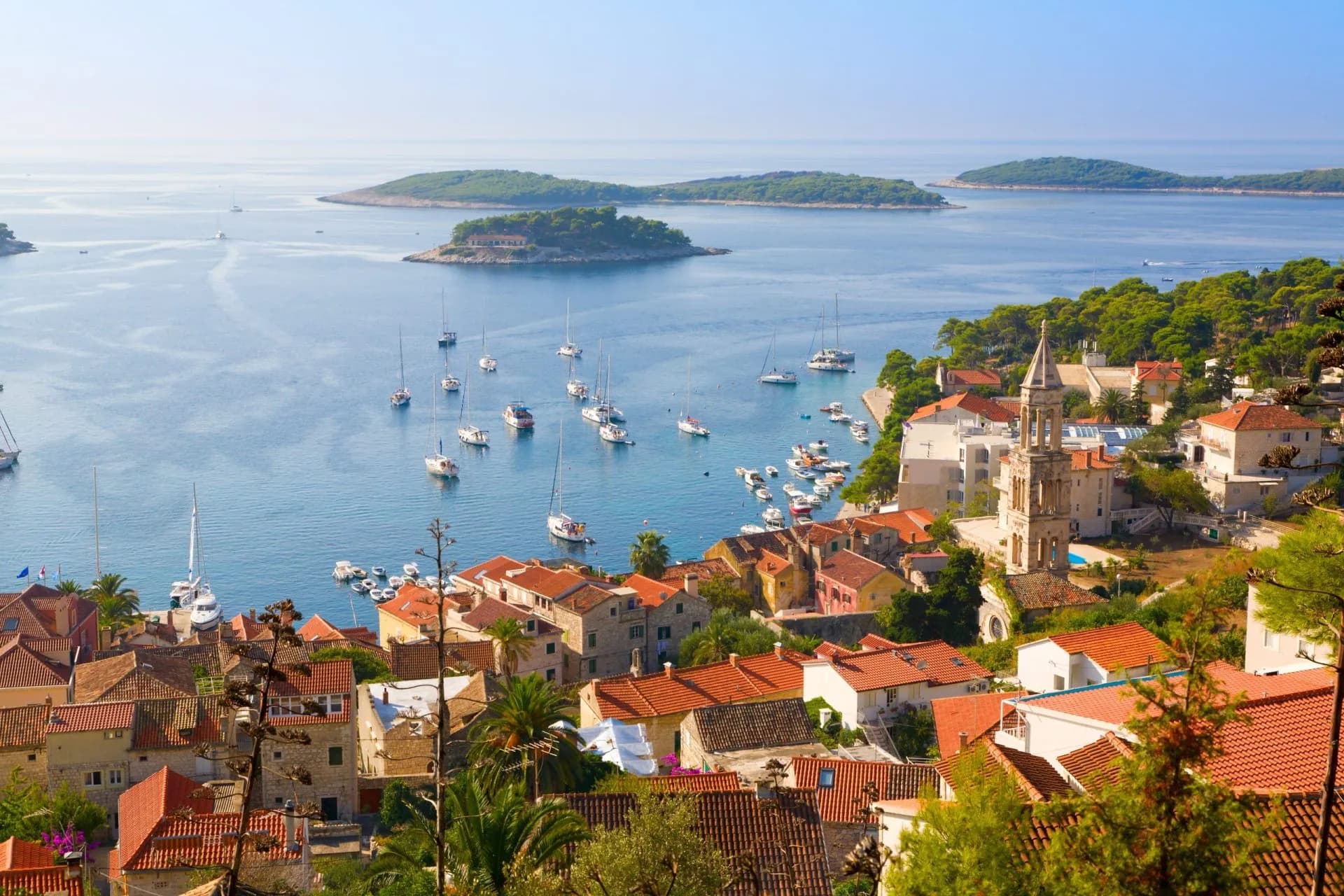Coastal town with terracotta roofs, bell tower, and many sailboats anchored in blue water.