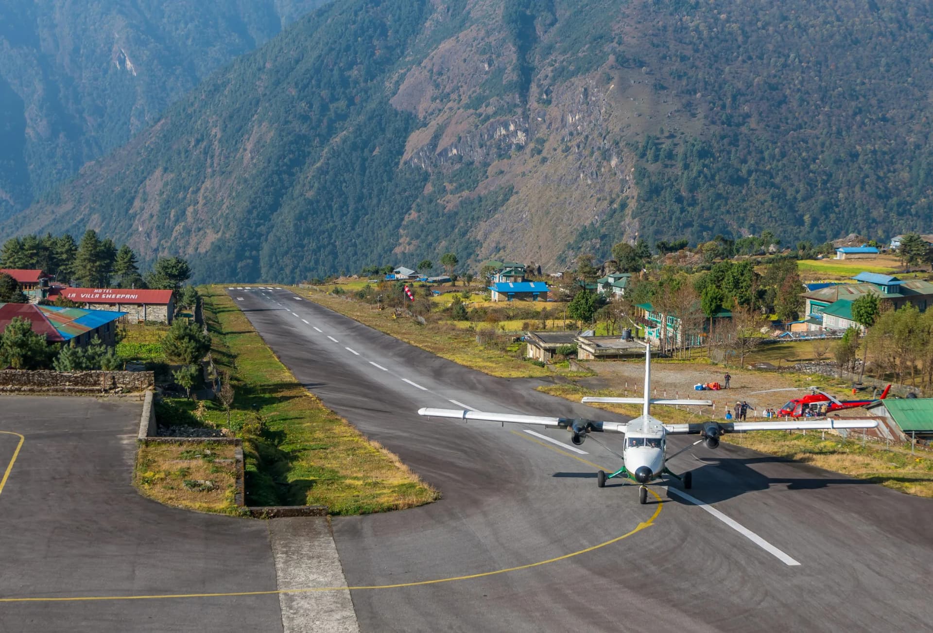 Small airplane on runway at Tenzing-Hillary Airport with steep mountains and village buildings.