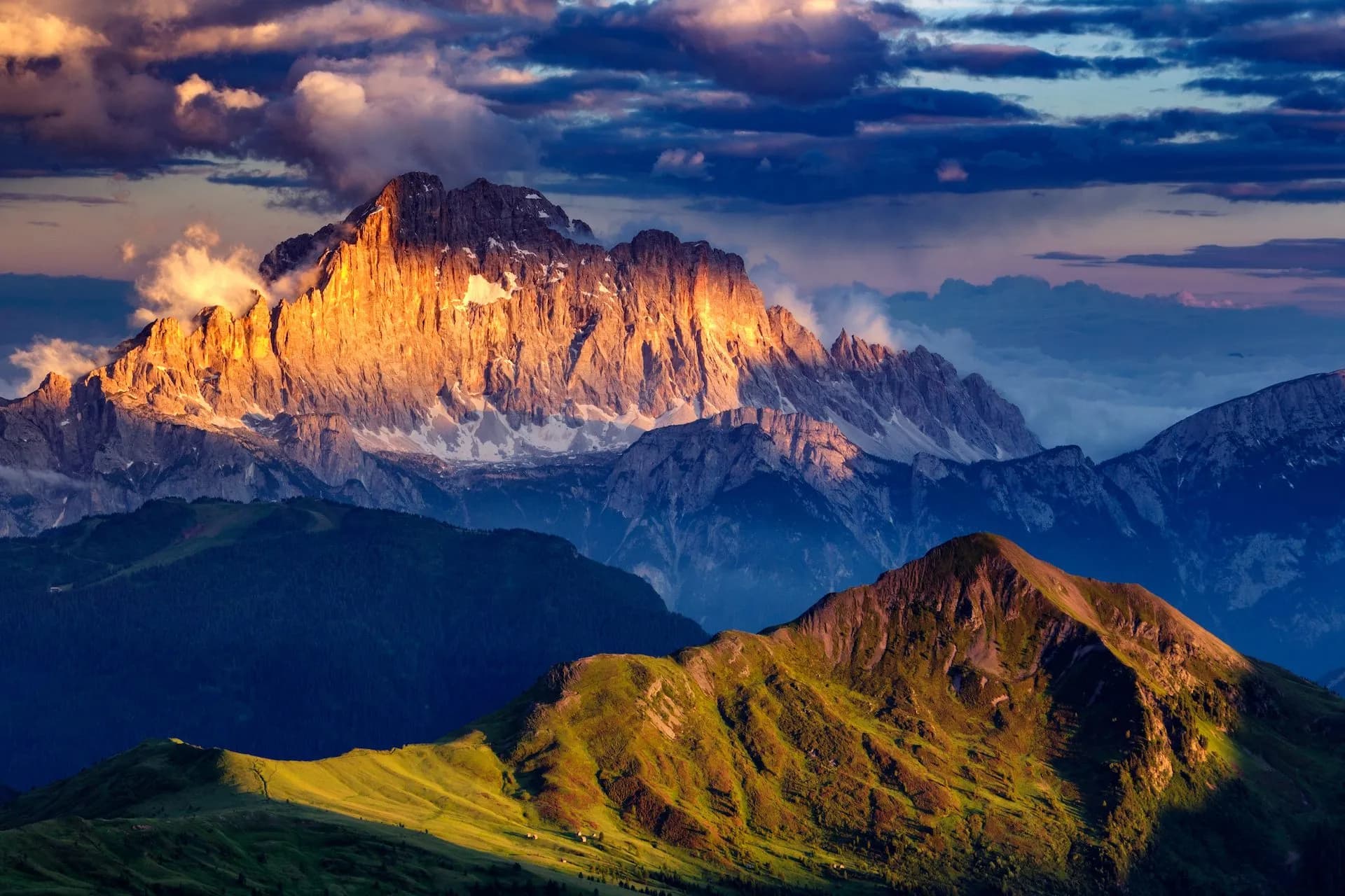 Rugged mountains illuminated by sunset light above green foothills under dramatic clouds, Monte Civetta.