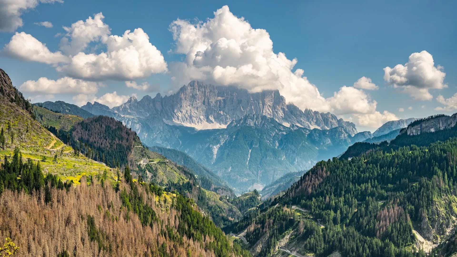 Rugged mountain peaks under clouds in the Dolomites, with green and brown forested slopes.