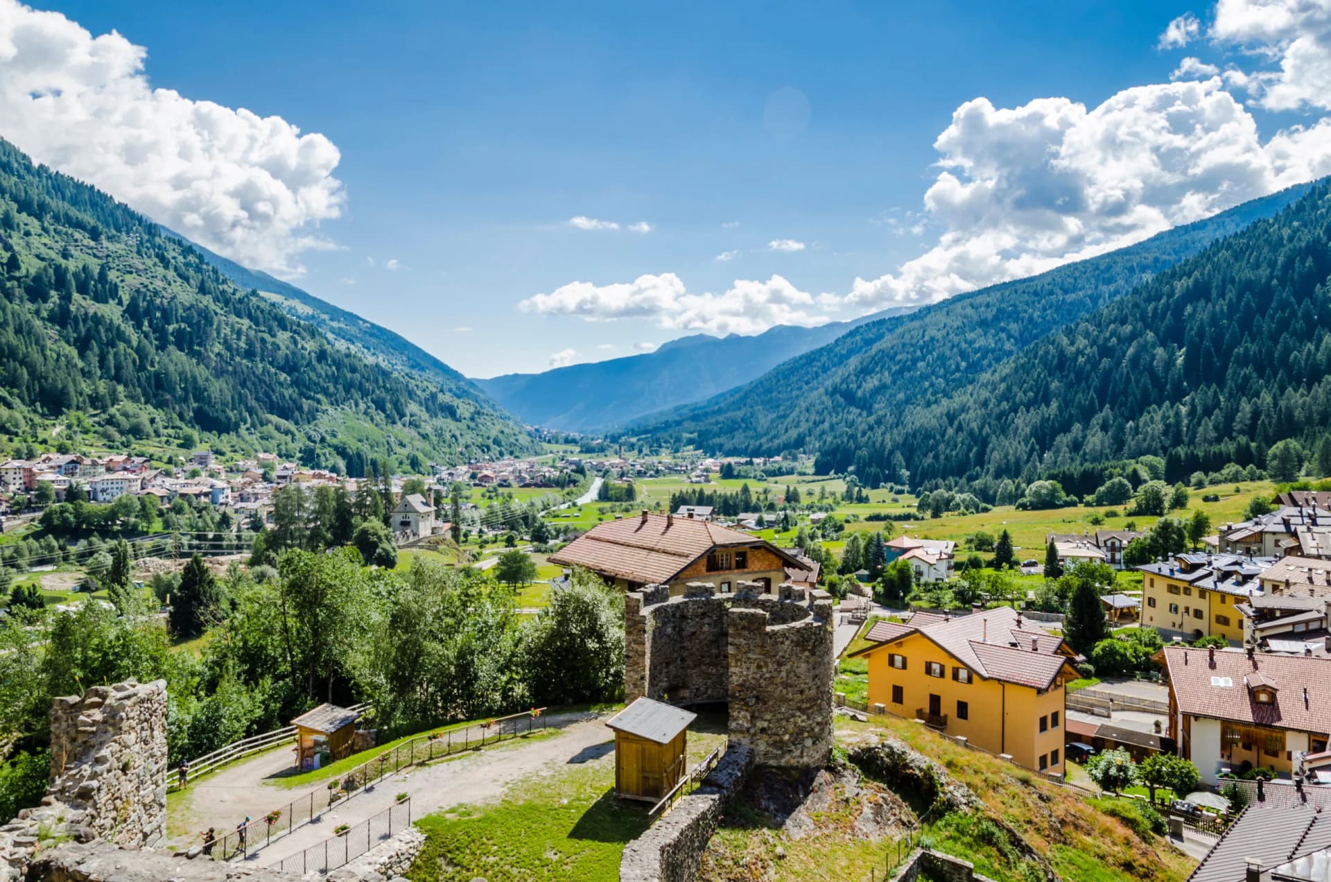 Alpine village valley with stone ruins, colorful houses, and forested mountains under blue sky.