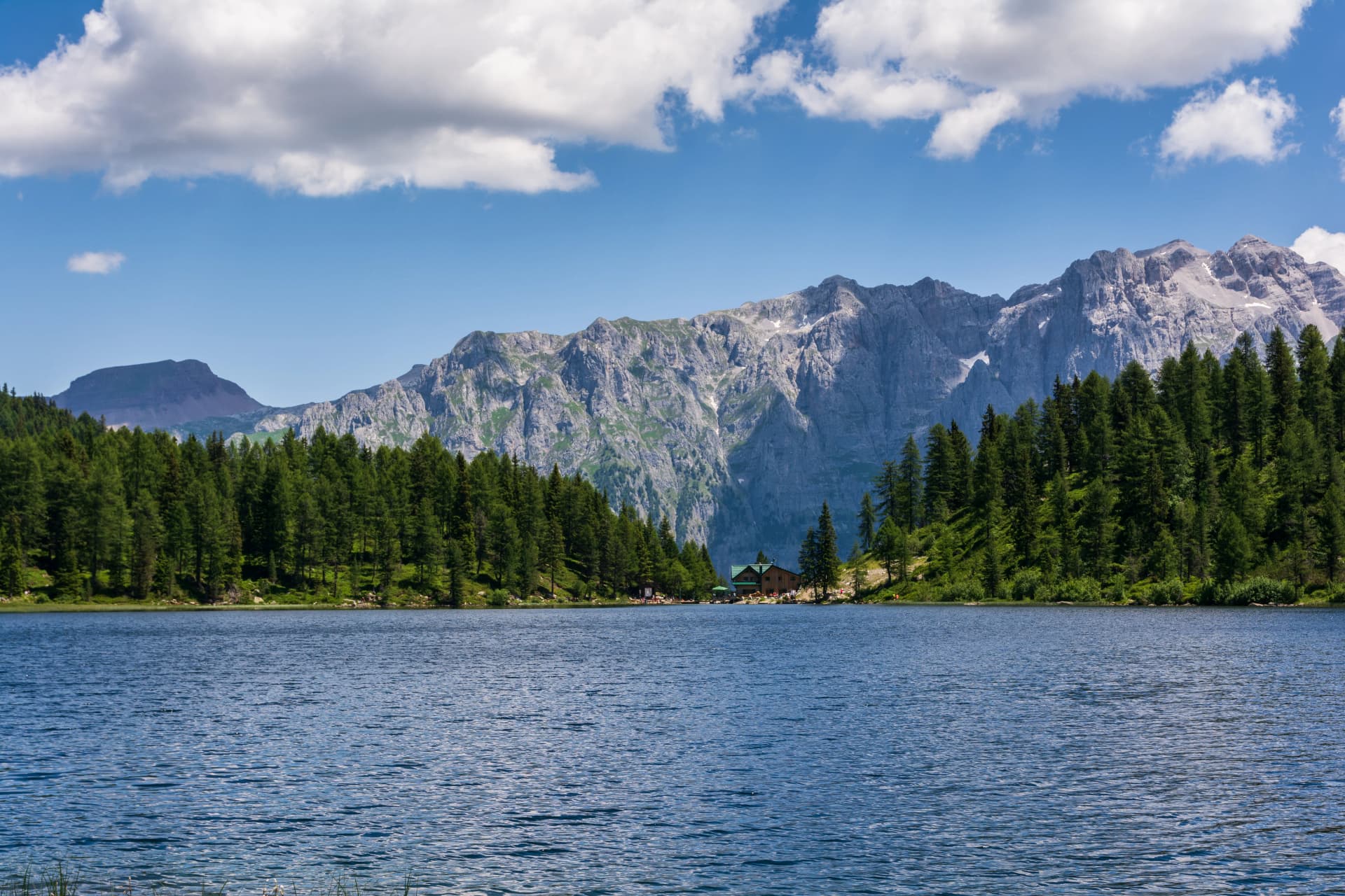 Alpine lake with dark blue water, pine forest, and rugged mountains under a blue sky with clouds.