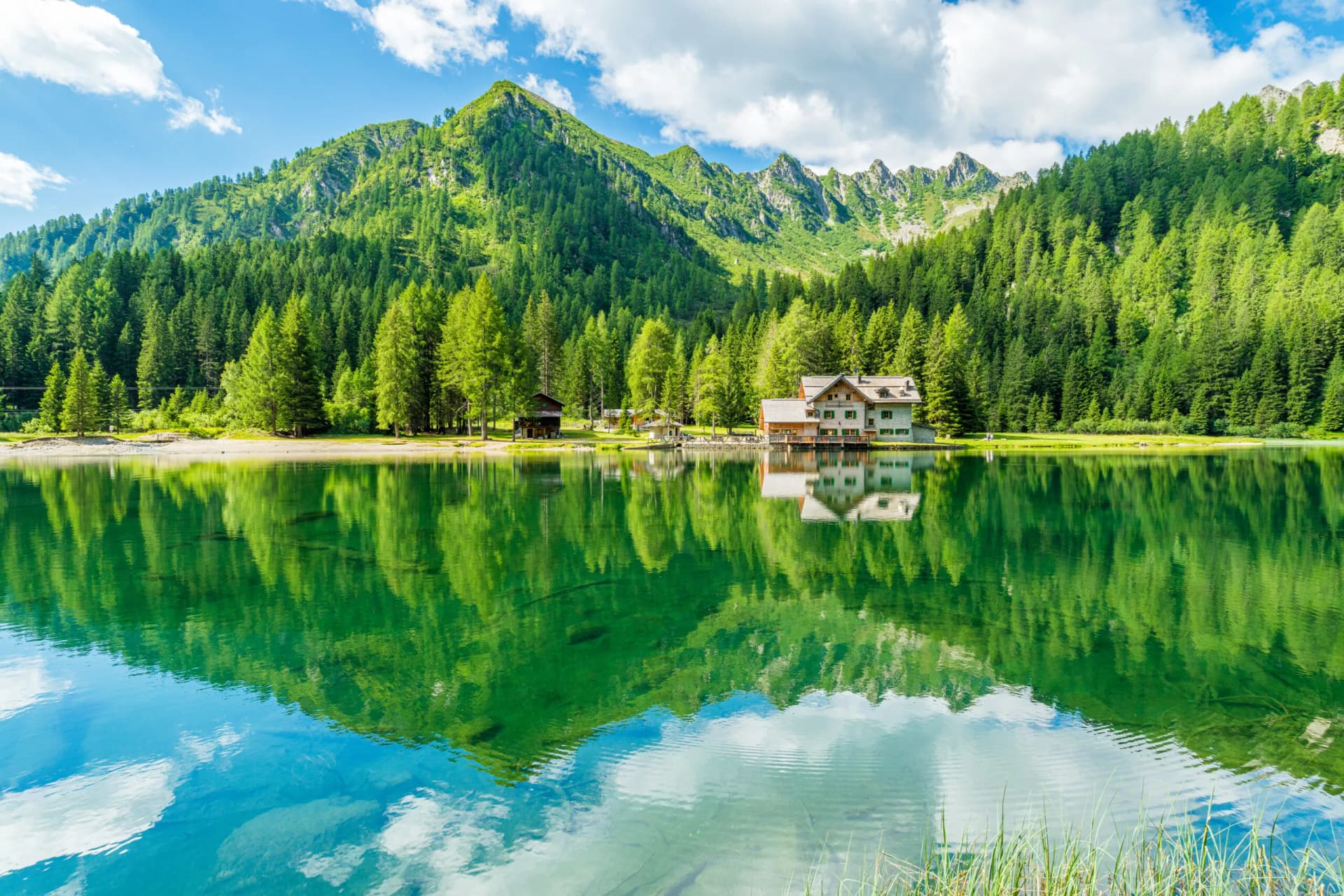 Alpine lake with Rifugio reflection, dense green forest, and rugged mountains under blue sky.