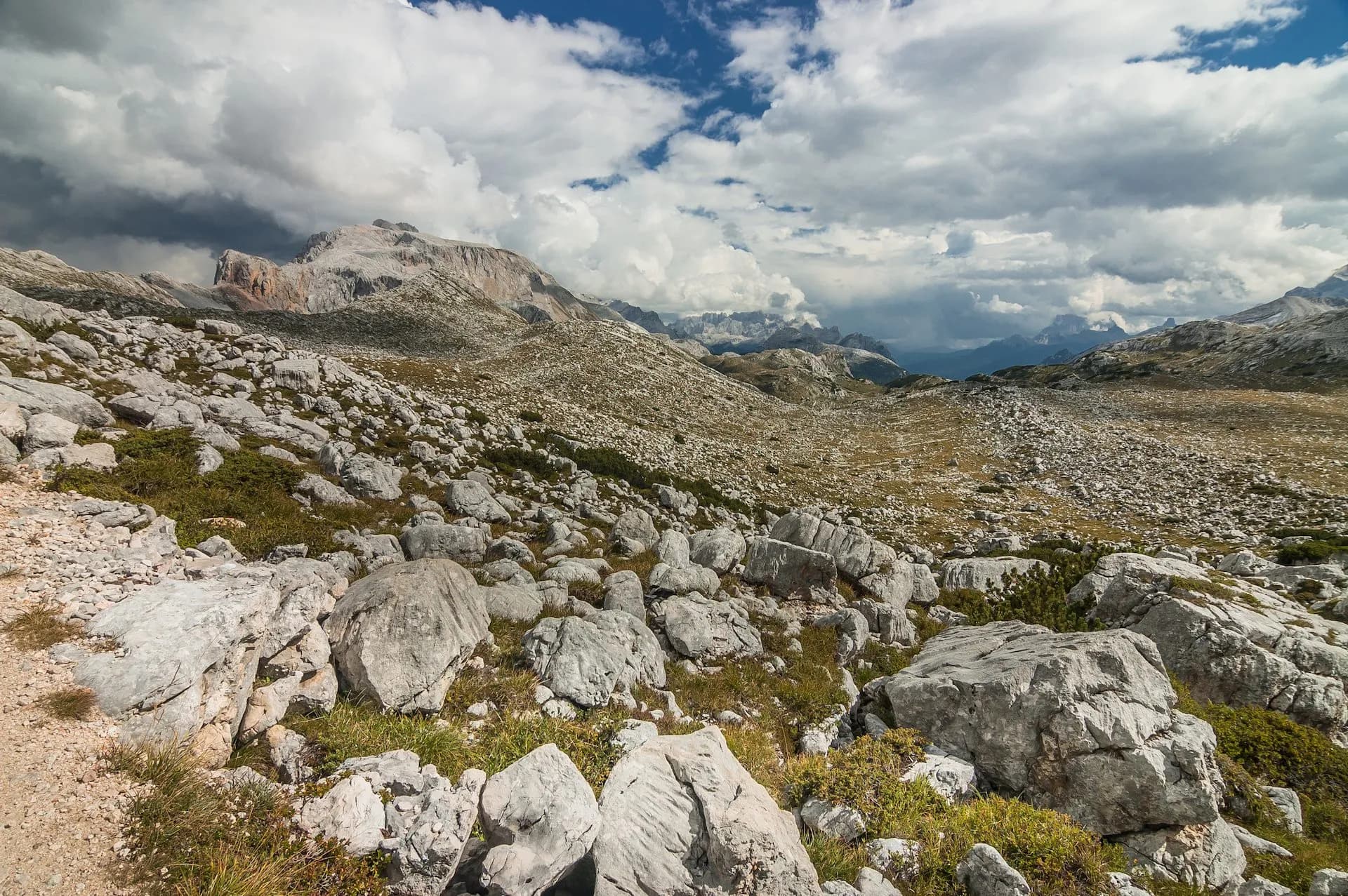 Rocky alpine terrain with large boulders, sparse grass, and distant mountains under a cloudy sky near Rifugio Biella.