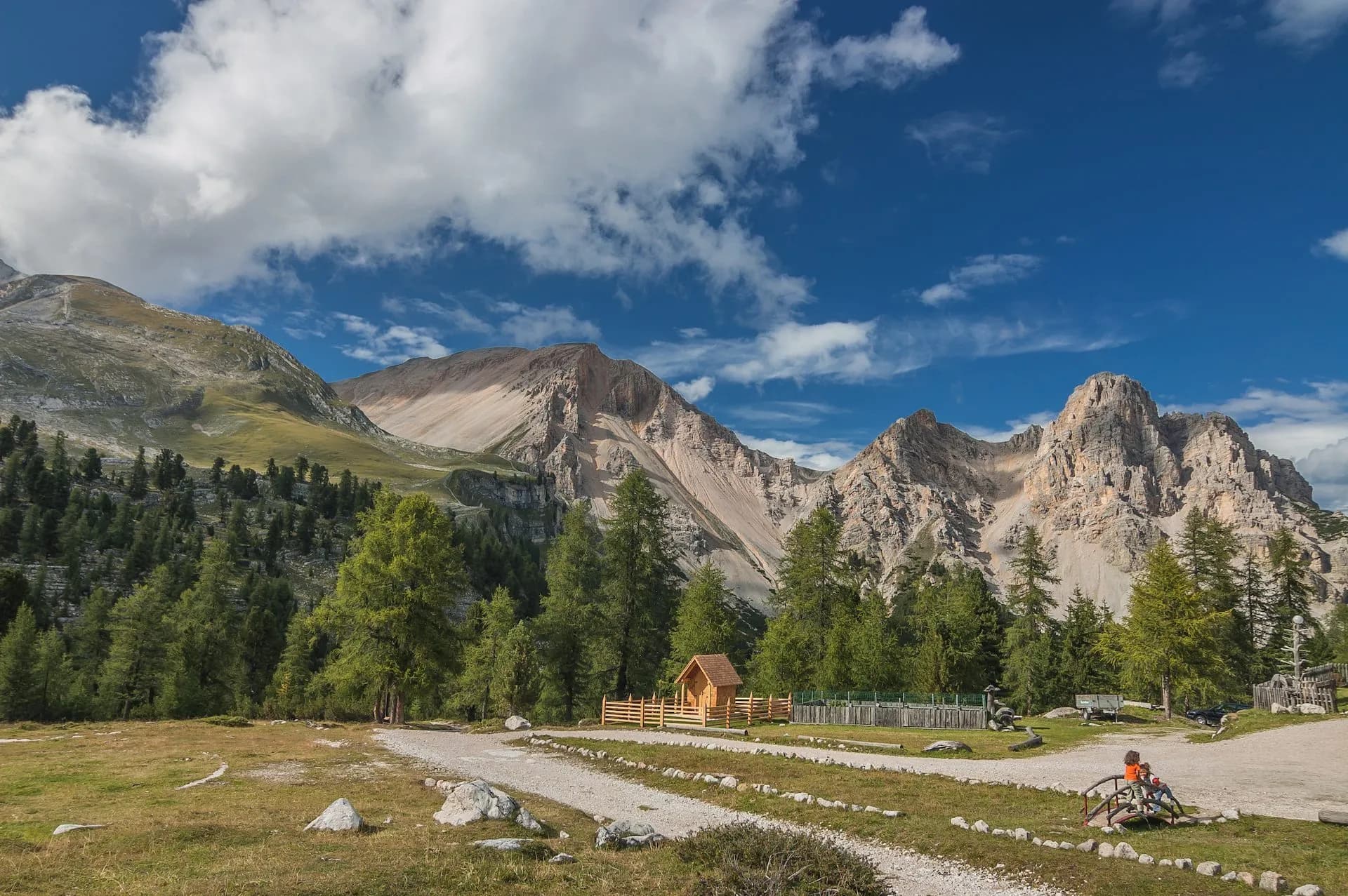 Dolomites mountains with rocky peaks, green forest, and a small wooden hut under a blue sky.