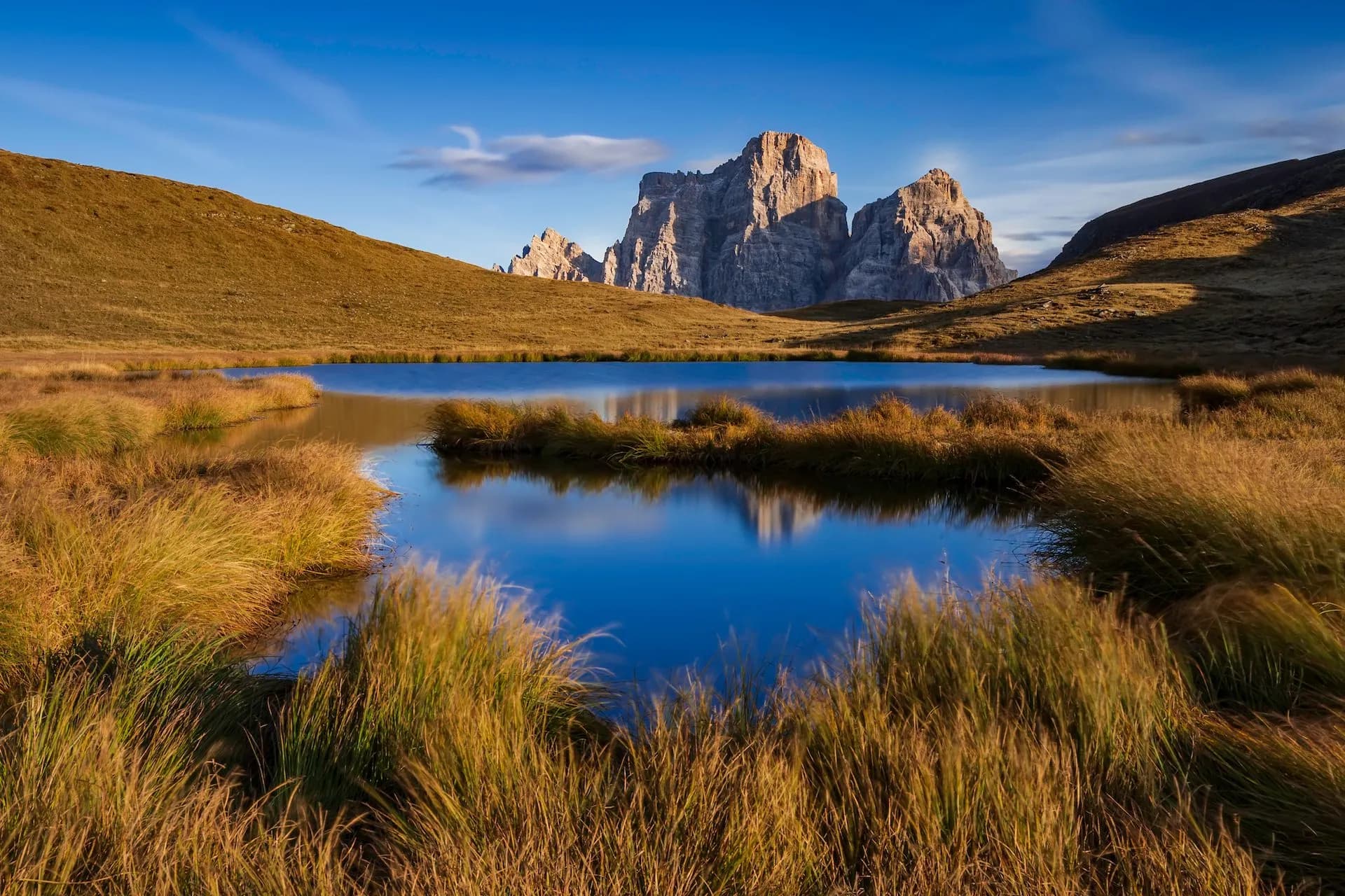 Mountain peaks reflected in a still alpine lake surrounded by golden autumn grass under a blue sky.