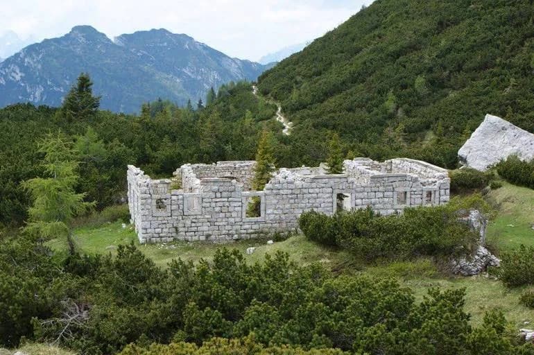 Ruins of stone structure in dense green mountainside with hiking path and distant peaks, Forte di Forcella Moschesi.