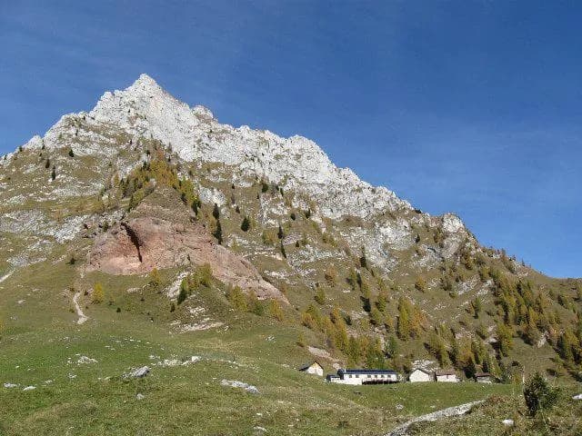 Rocky mountain peak above alpine meadow with small buildings and scattered autumn trees under blue sky.