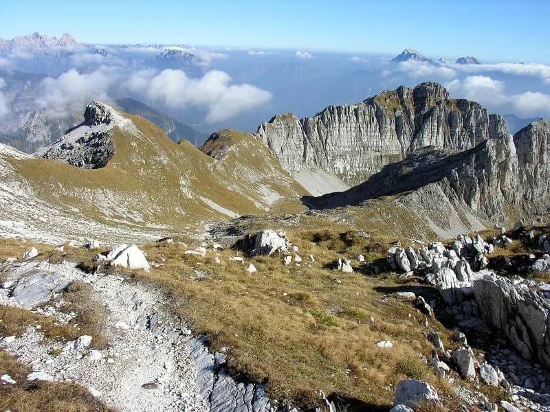 Rocky alpine terrain with steep cliffs, dry grass, and distant mountain peaks above clouds